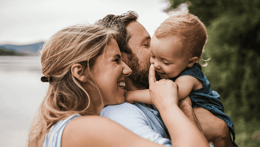 Сім'я на природі, батьки обіймають дитину, батько цілує її в щоку, а мати усміхається. Всі троє разом, виражаючи любов і прив’язаність.