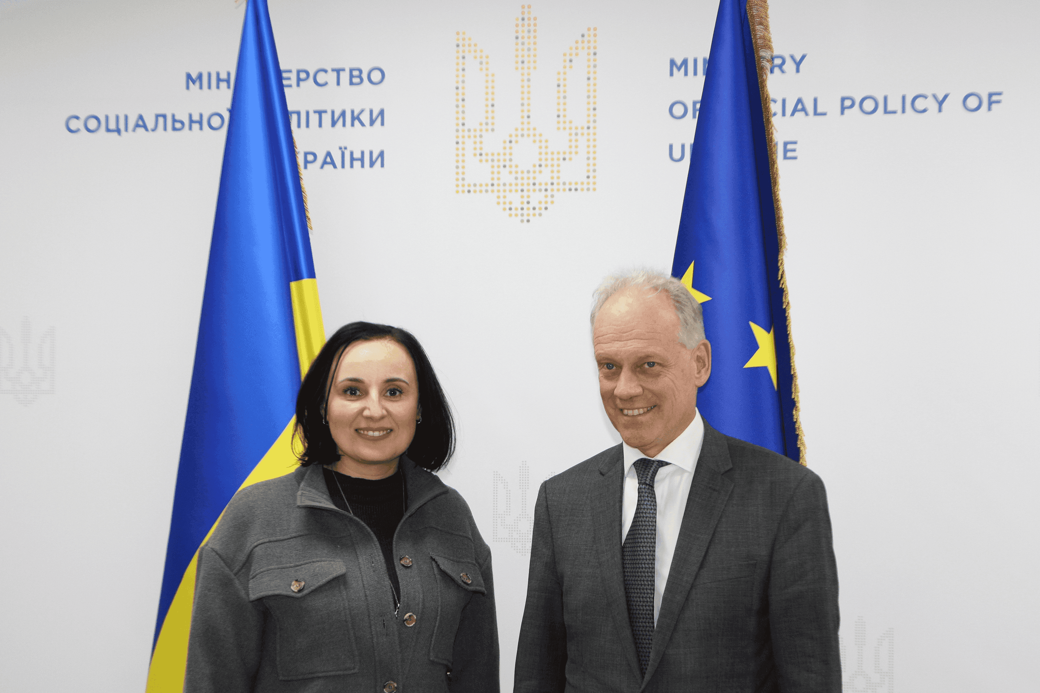 Міністр соціальної політики України Оксана Жолнович та генеральний директор Генерального директорату Європейської Комісії із питань розширення та Східного сусідства (DG ENEST) Герт Купман фотографуються на фоні прапорів України  та ЄС
