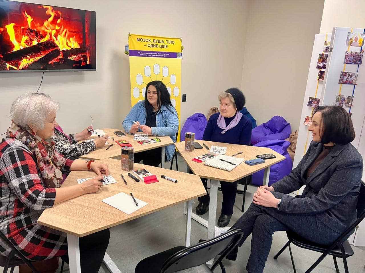 Чотири жінки сидять за столом у кімнаті. Вони працюють разом над творчим заняттям, на столі розкладені різні матеріали для рукоділля. На фоні видно телевізор, на якому зображене полум'я каміна. Одна з жінок розповідає