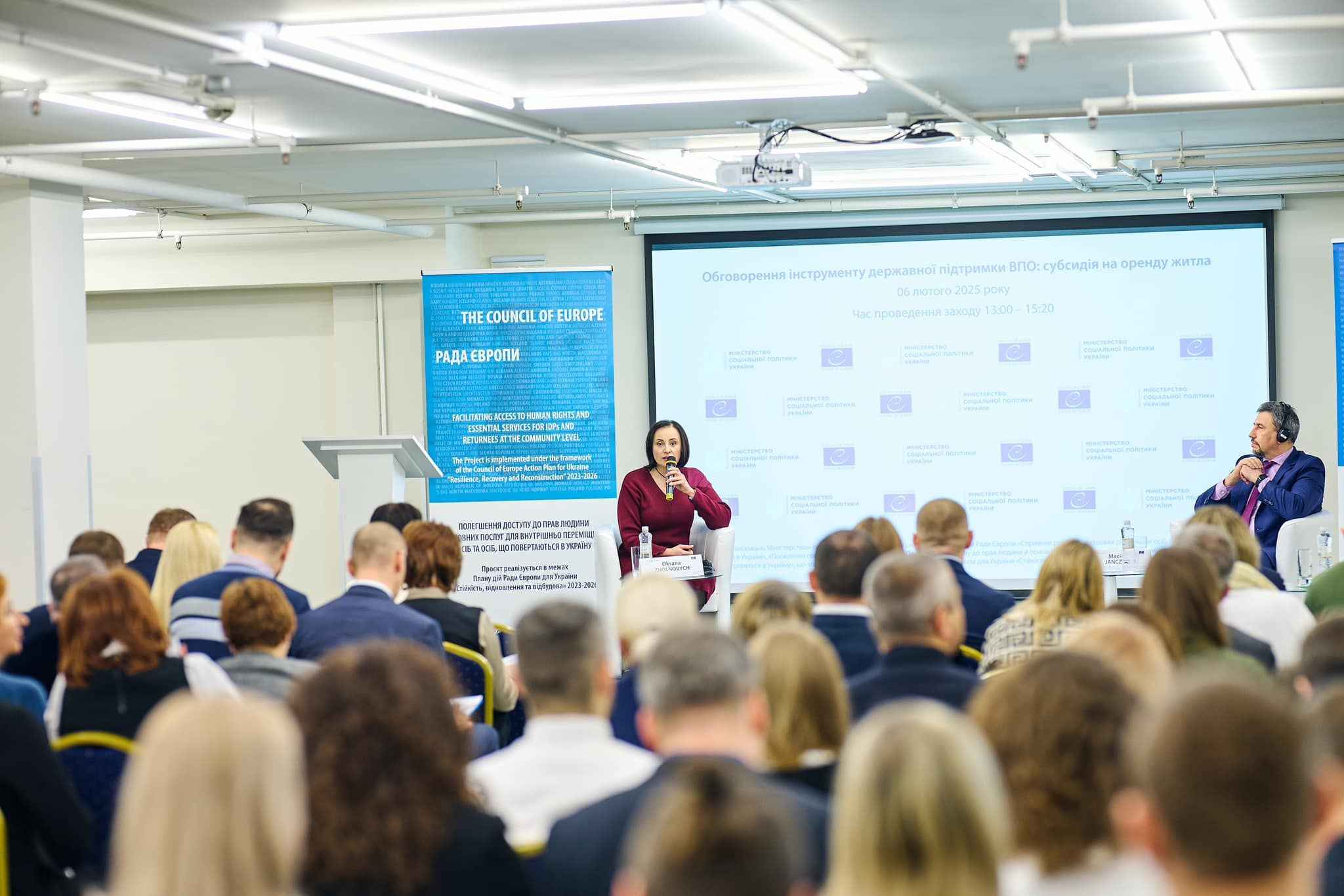 На зображенні велика аудиторія під час конференції або семінару. На екрані презентація