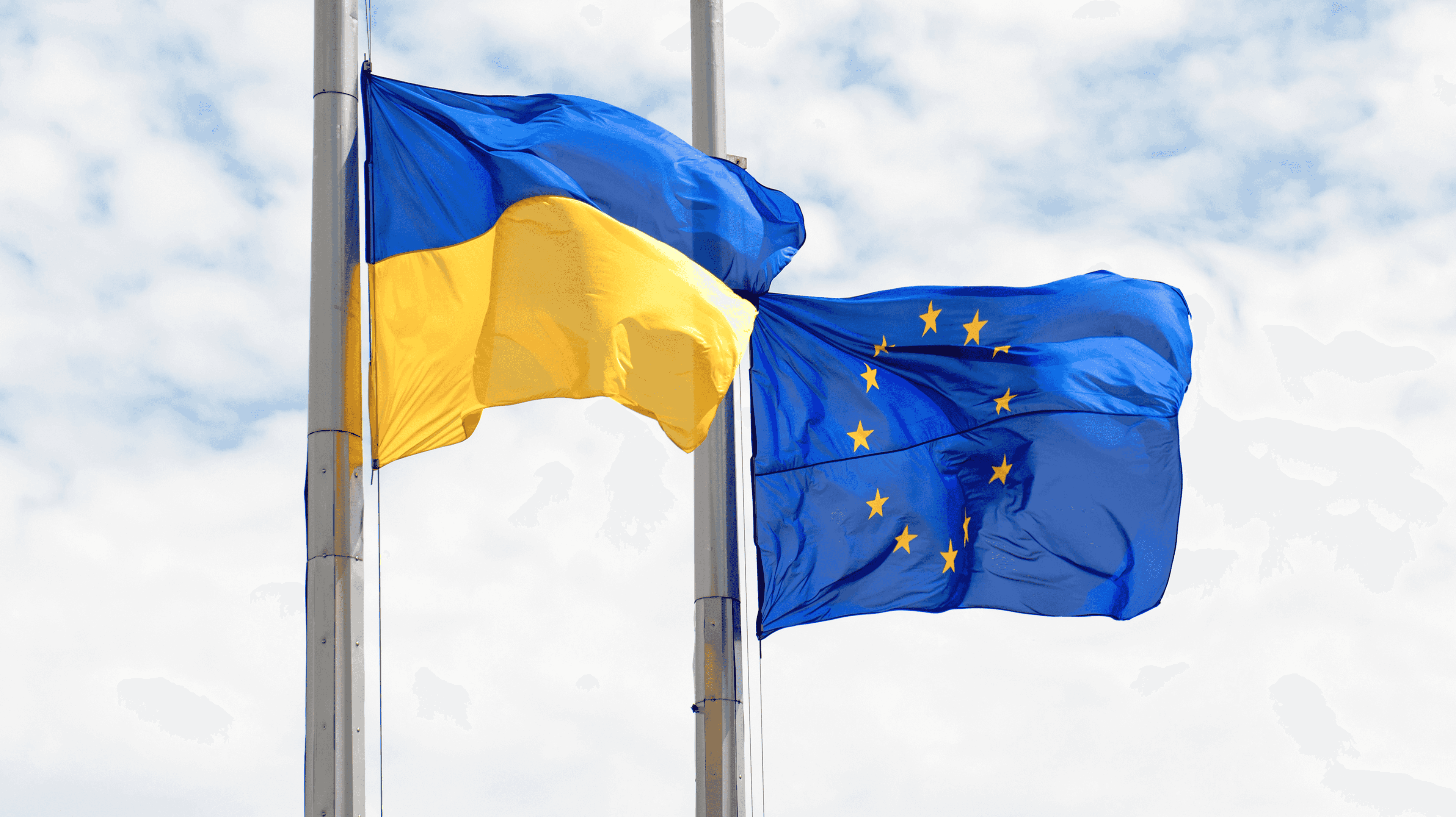На зображенні два прапори, що майорять на флагштоках: зліва – прапор України (жовто-блакитний), а справа – прапор Європейського Союзу (синій із жовтими зірками). Фон – хмарне небо.