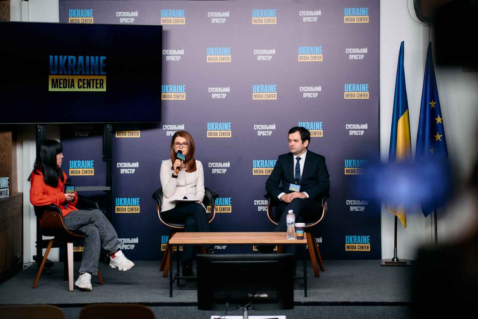 На фото жінки та чоловік сидять на фоні банеру під час заходу