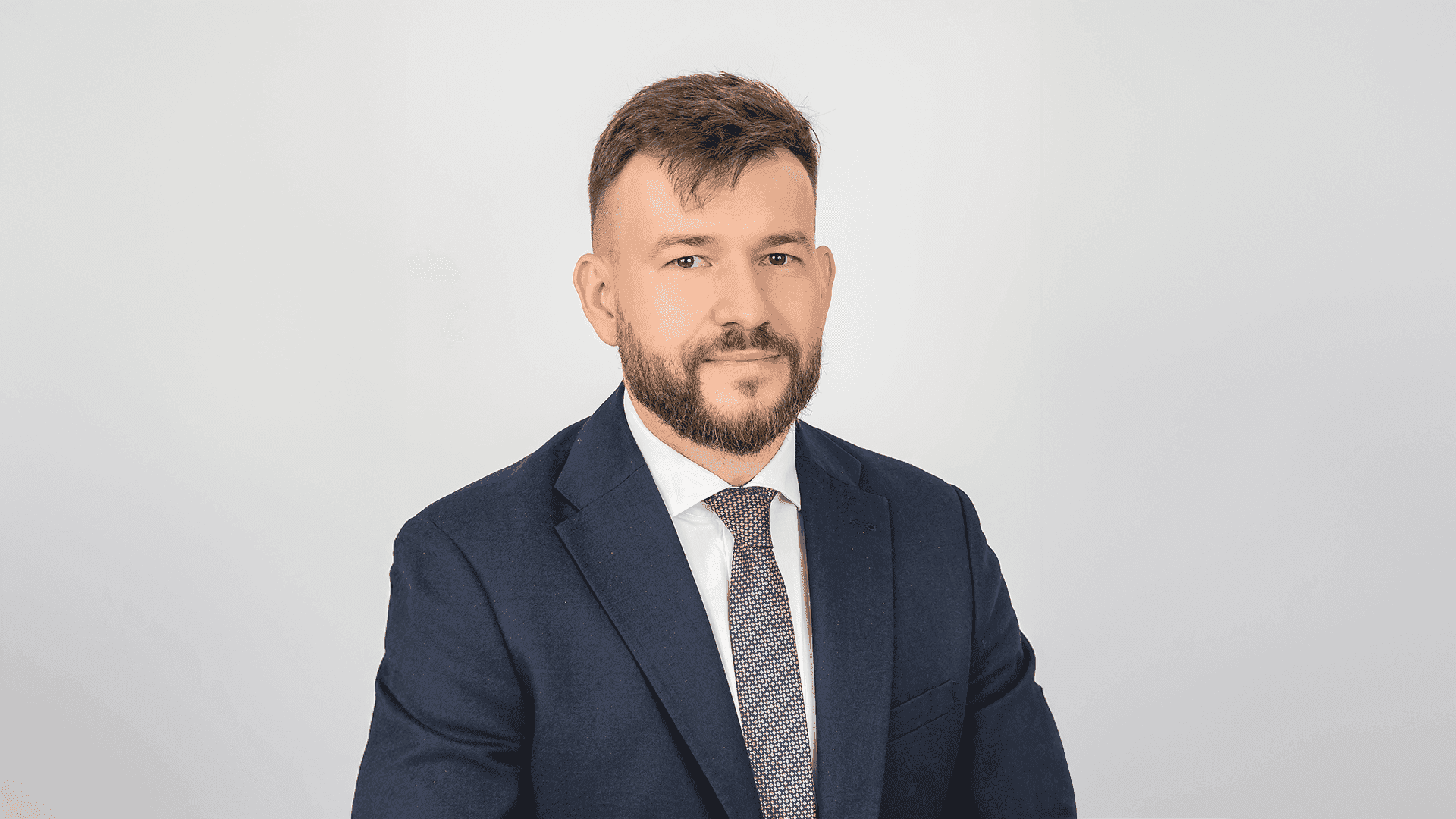 Чоловік у темно-синьому костюмі, білій сорочці та краватці з дрібним візерунком. Має коротку стрижку та бороду.