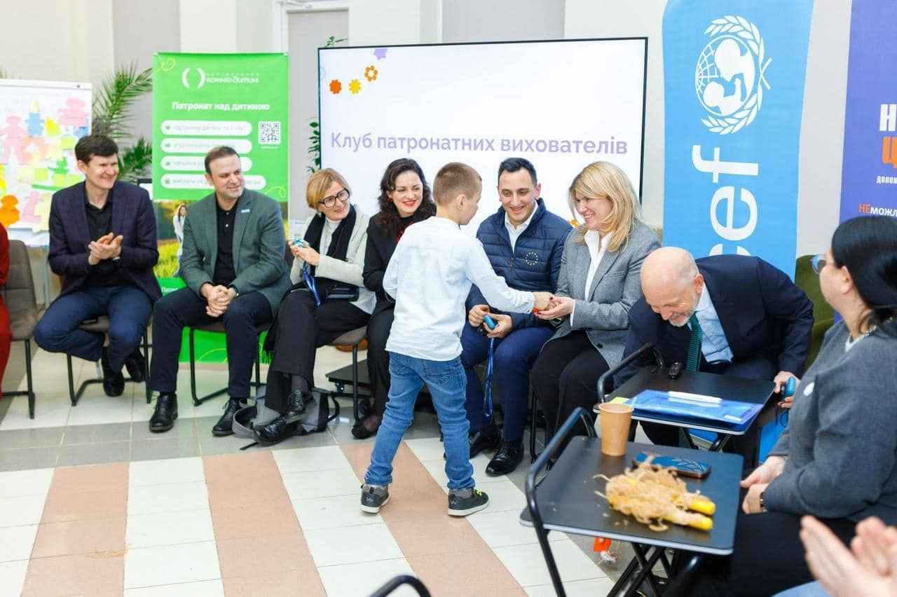 Дитина, одягнена в білу сорочку, тисне руку одному з дорослих під час заходу. Інші учасники сидять поруч і спостерігають за подією. На фоні видно логотипи UNICEF та інших організацій.
