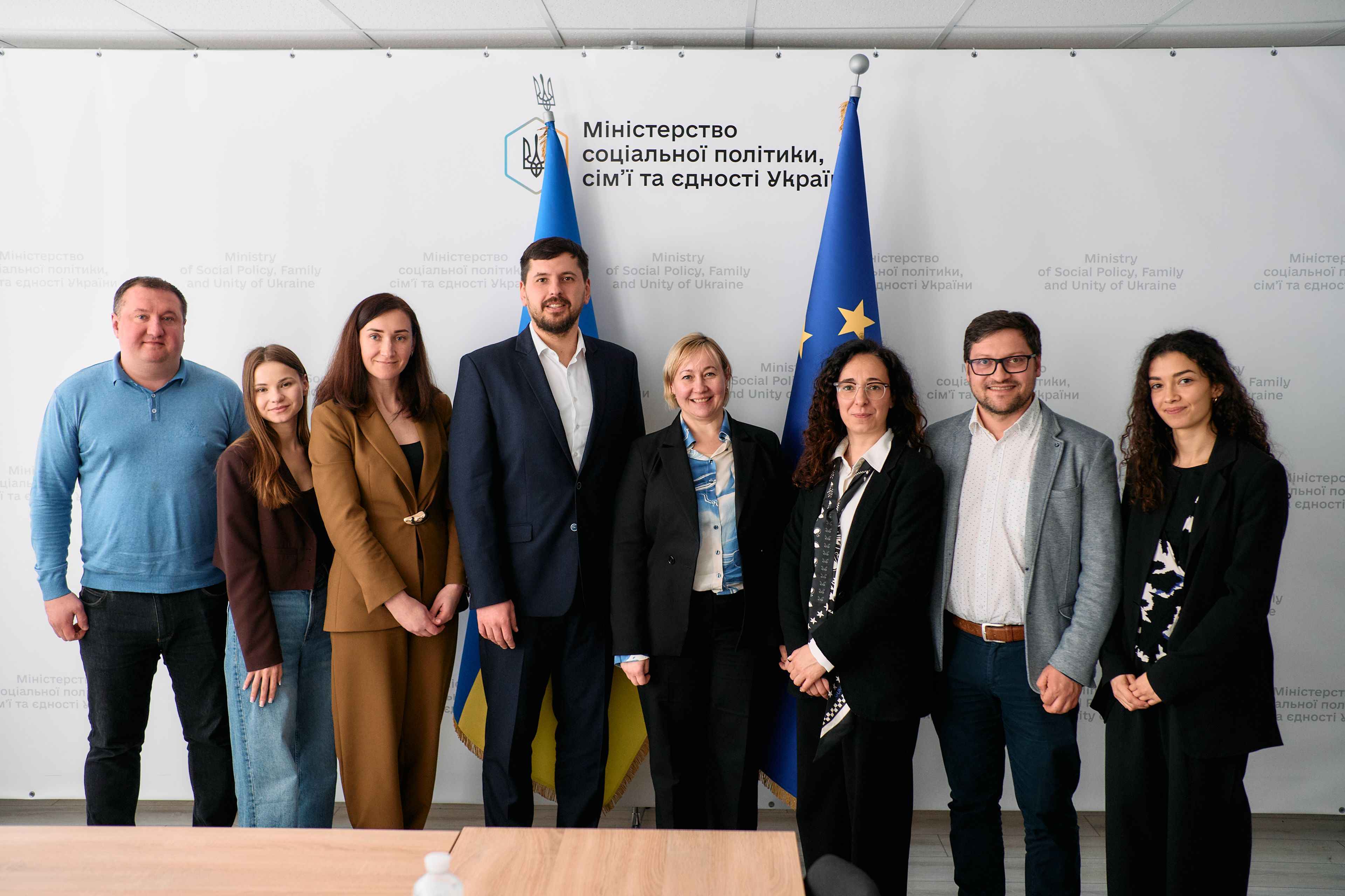 На фото чоловіки та жінки стоять на фоні прапорів України та ЄС