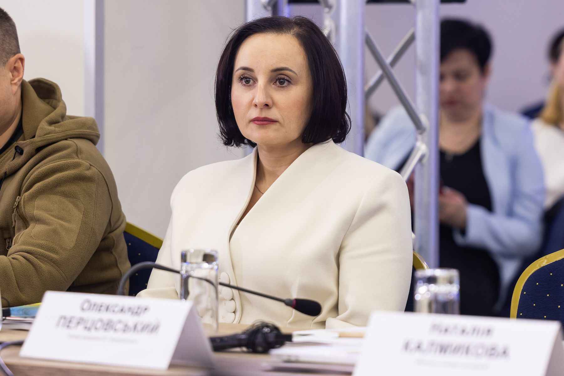 На зображенні жінка, Міністерка Оксана Жолнович, з коротким чорним волоссям у білому костюмі сидить за столом на офіційному заході. Вона має серйозний вираз обличчя. Перед нею мікрофон, склянка води та документи