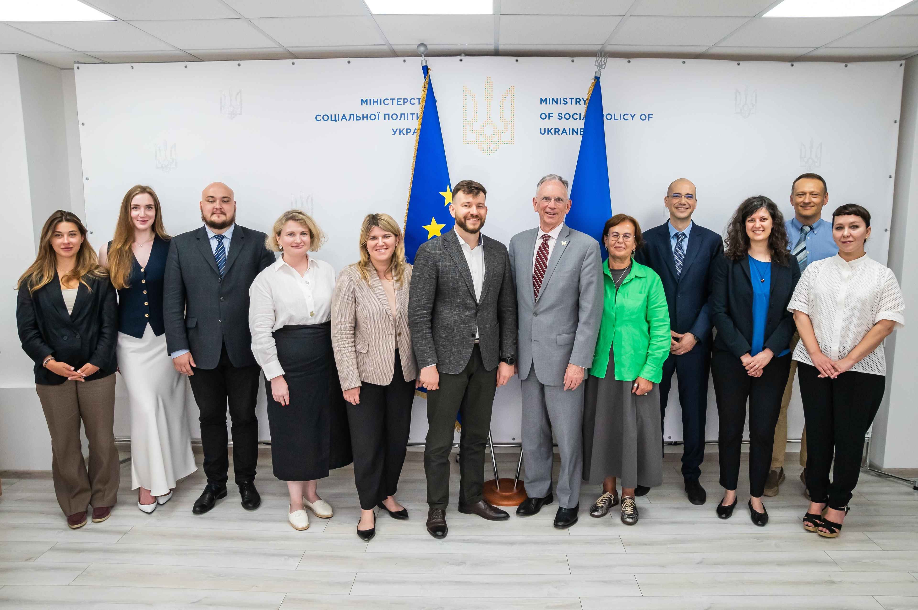 На фото чоловіки та жінки стоять на фоні прапорів ЄС та України