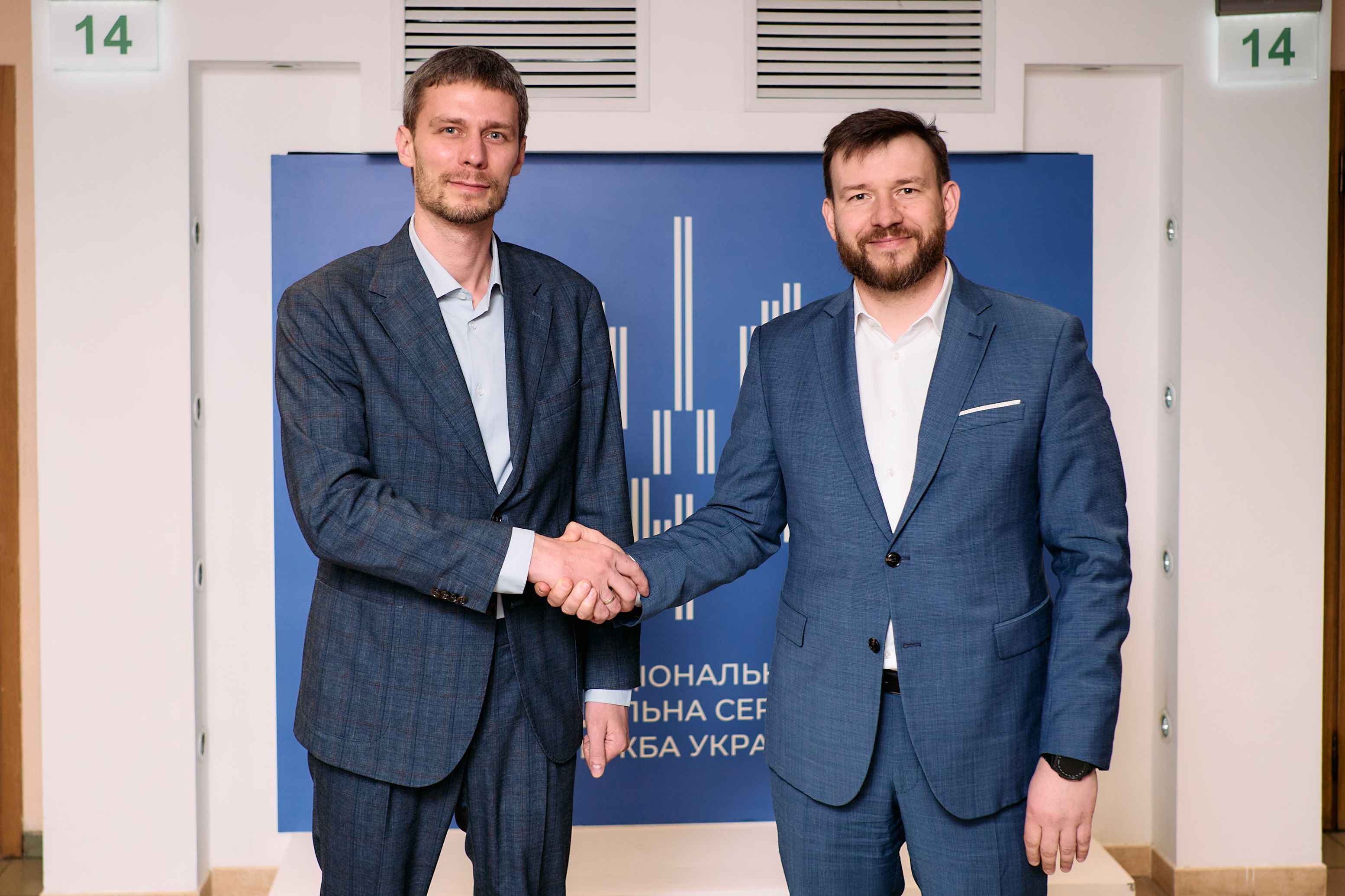 На фото чоловіки вітаються за руку на фоні банеру Нацсоцслужби