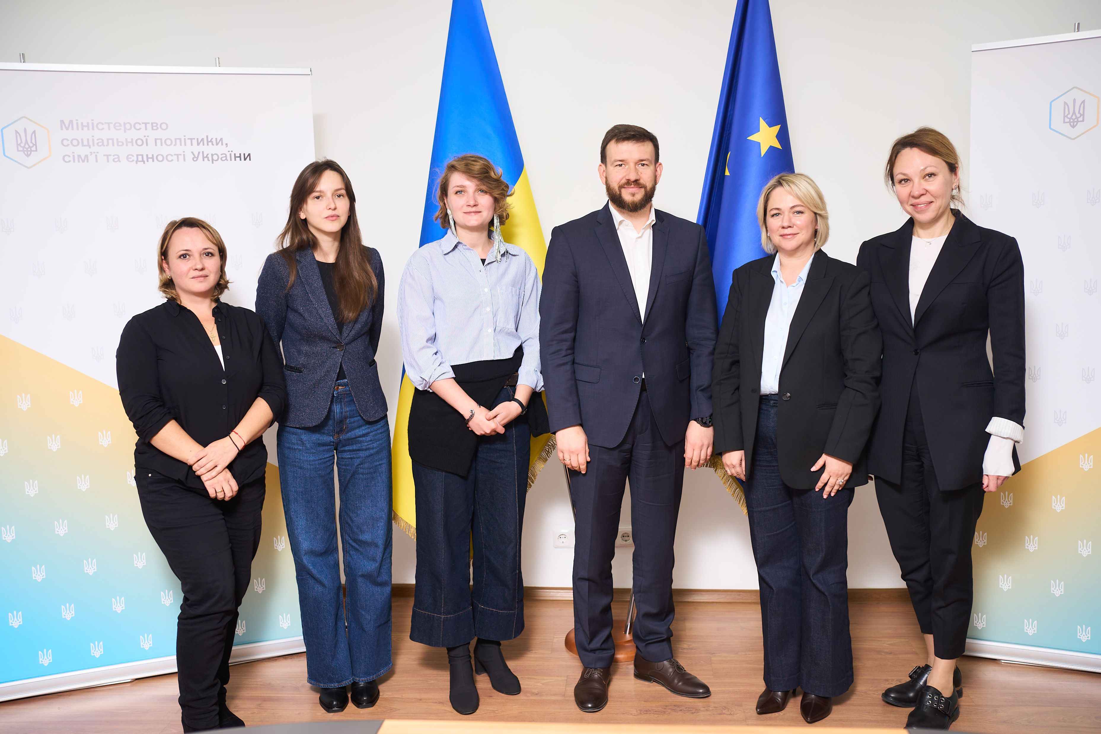 НА фото єінки та чоловік стоять на фоні прапорів України та ЄС