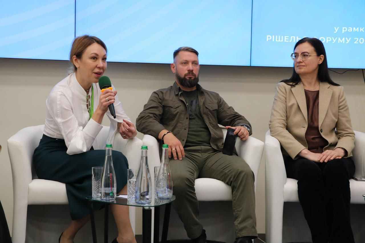 На фото жінки та чоловік сидять у президії під час заходу