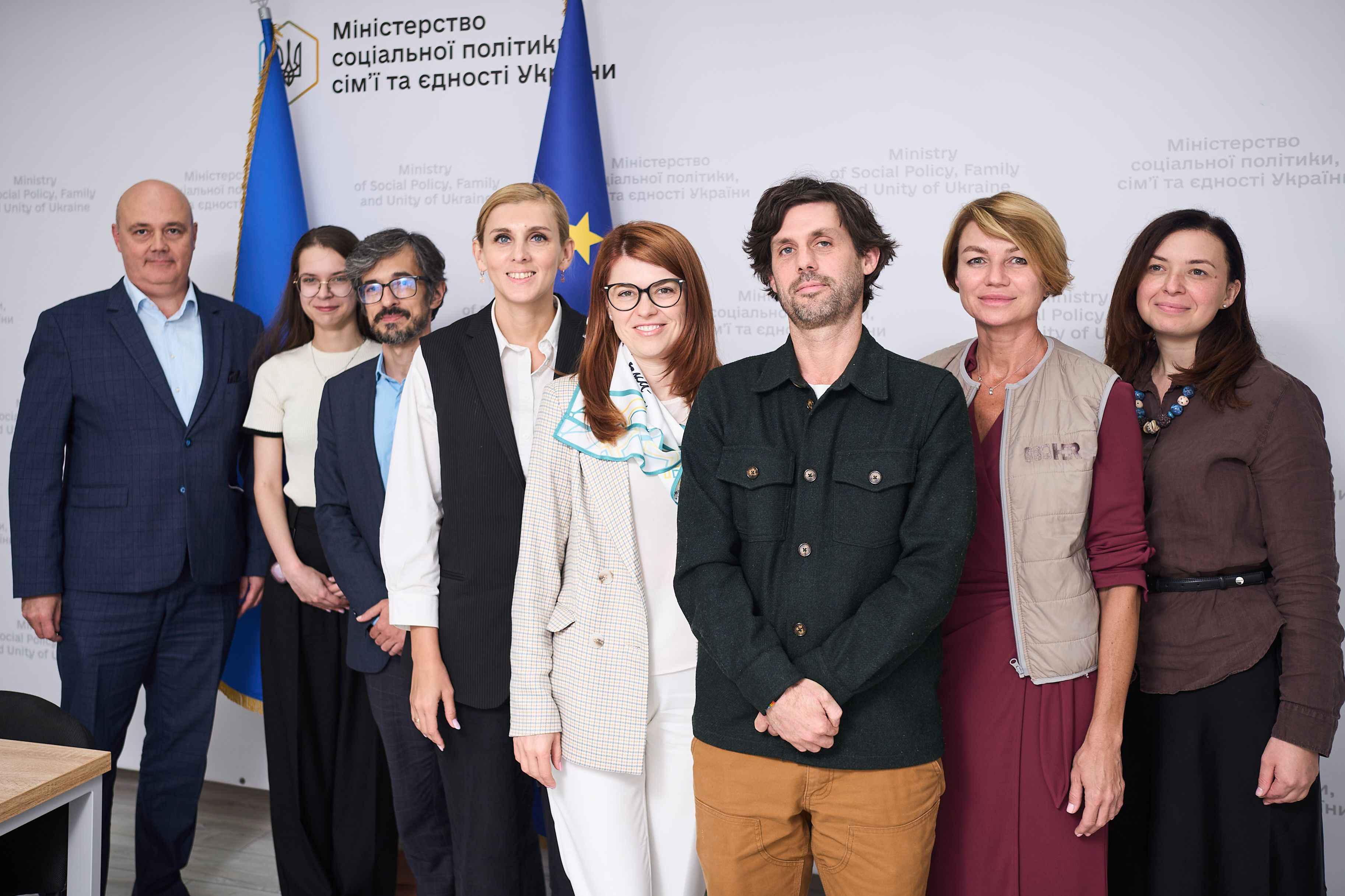 На фото жінки та чоловіки стоять на фоні прапорів України та ЄС