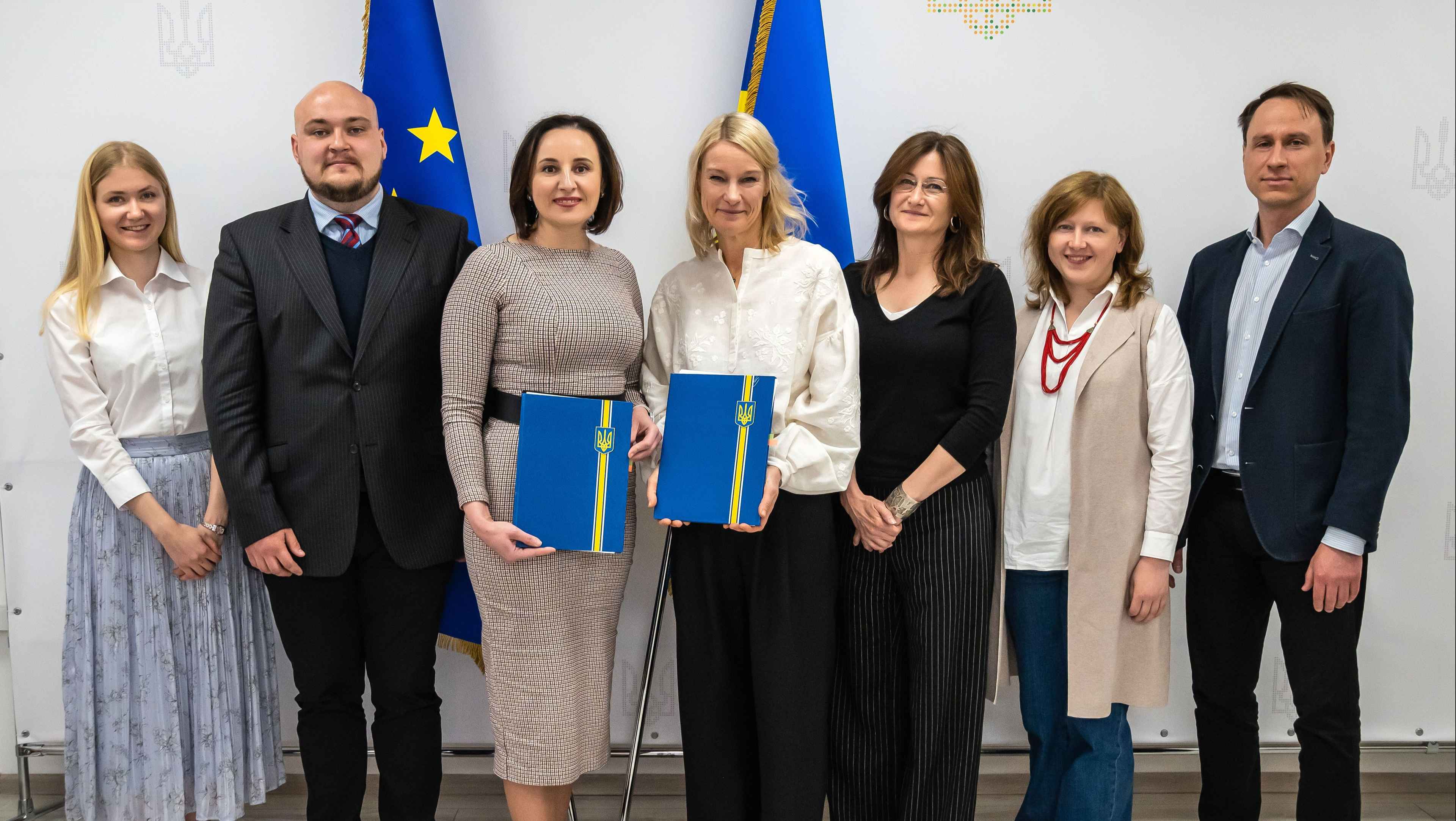 На фото група людей після підписання документів. Посміхаються і тримають в руках папки синього кольору. 