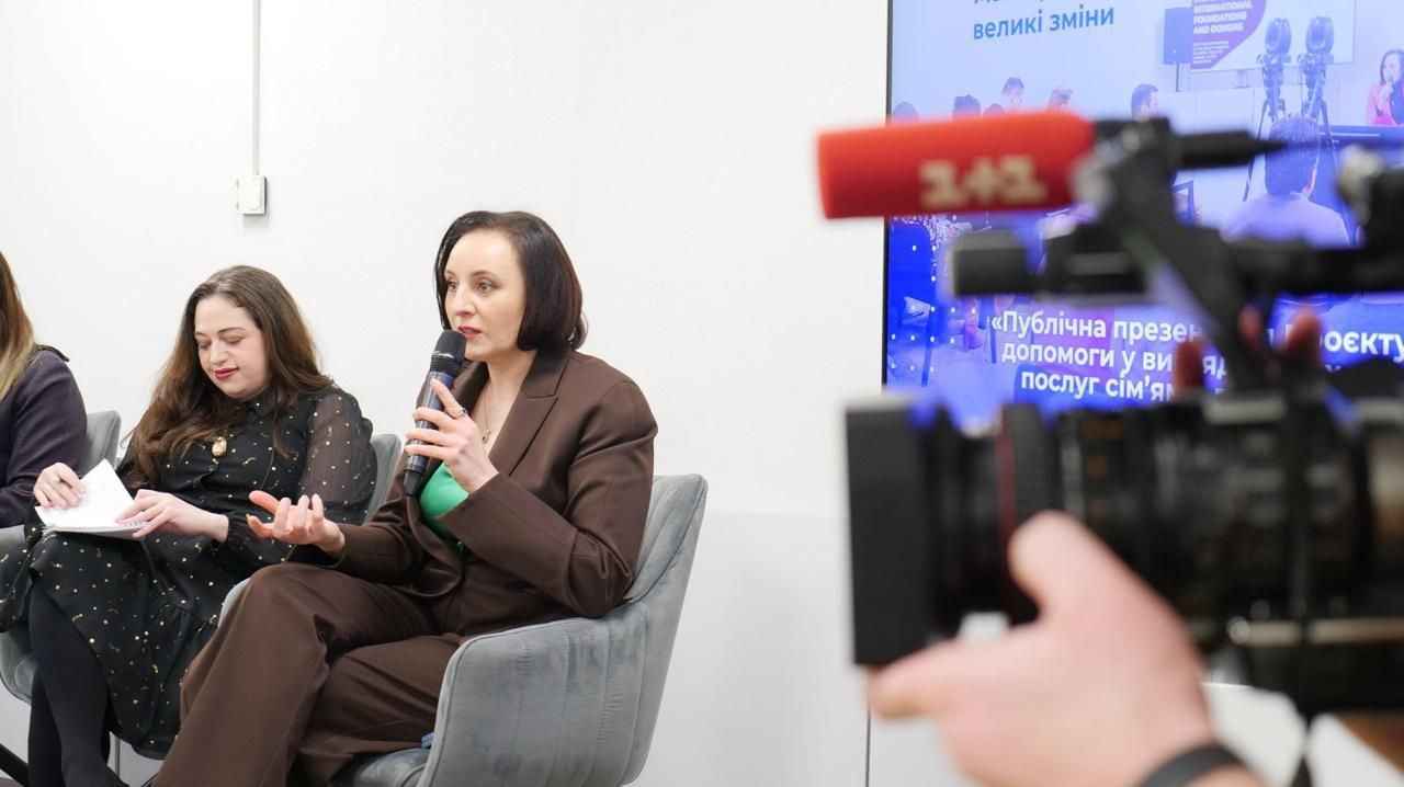 На зображенні зображена публічна подія, ймовірно, пресконференція або презентація. Міністерка Оксана Жолнович в коричневому костюмі з мікрофоном в руці говорить, а поруч з нею сидить інша жінка, яка робить нотатки