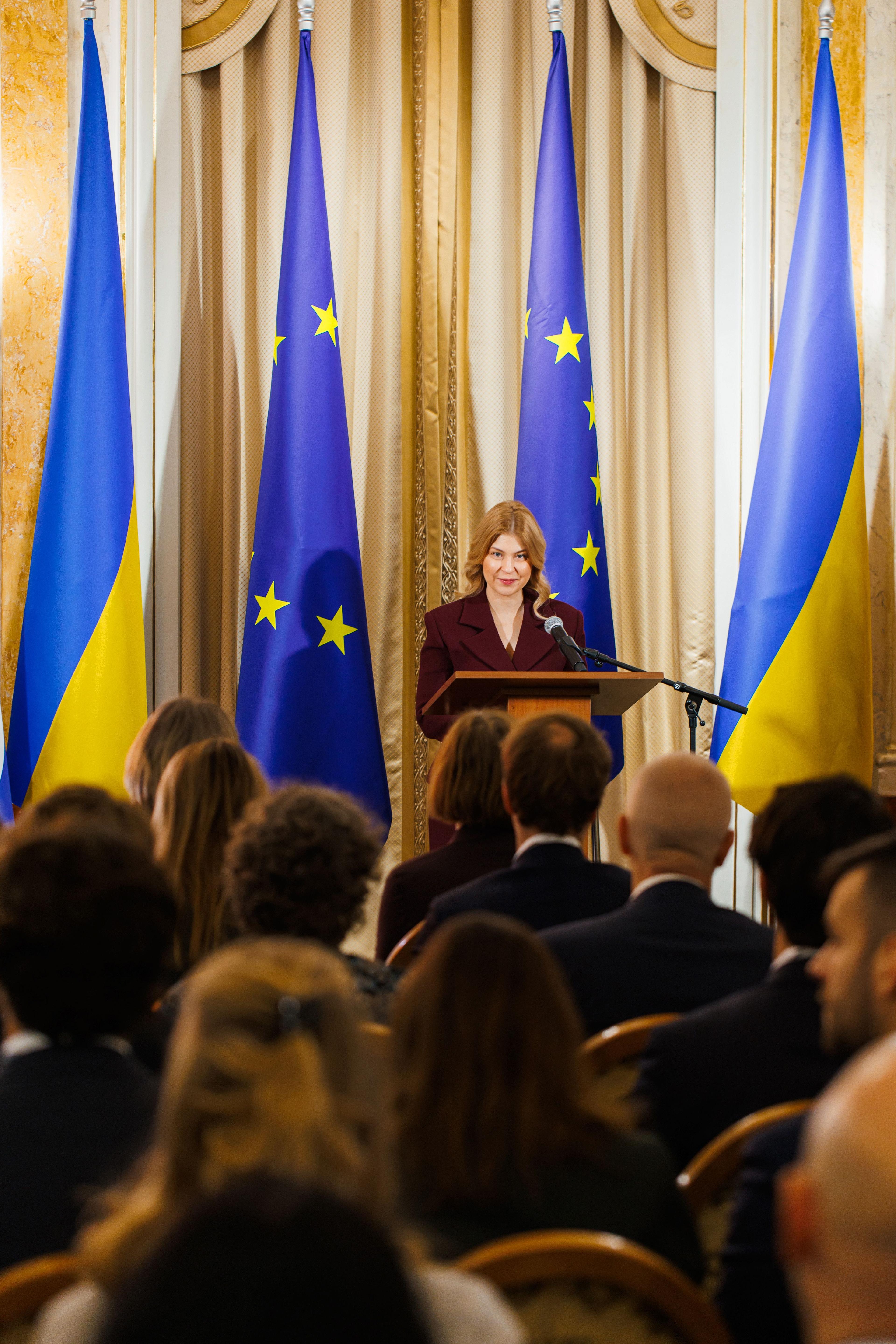 На зображенні жінка в бордовому костюмі щось доповідає. На задньому фоні пропори.