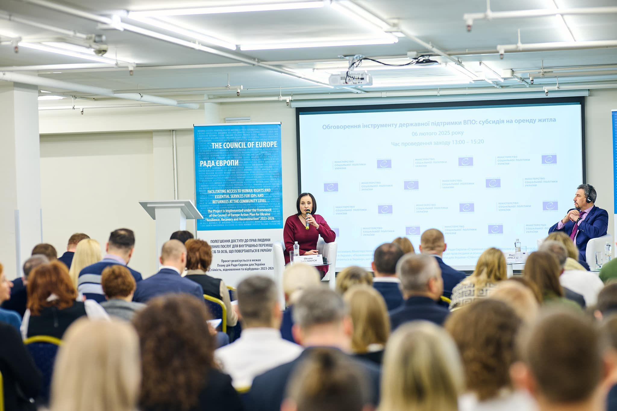 На зображенні велика аудиторія під час конференції або семінару. На екрані презентація