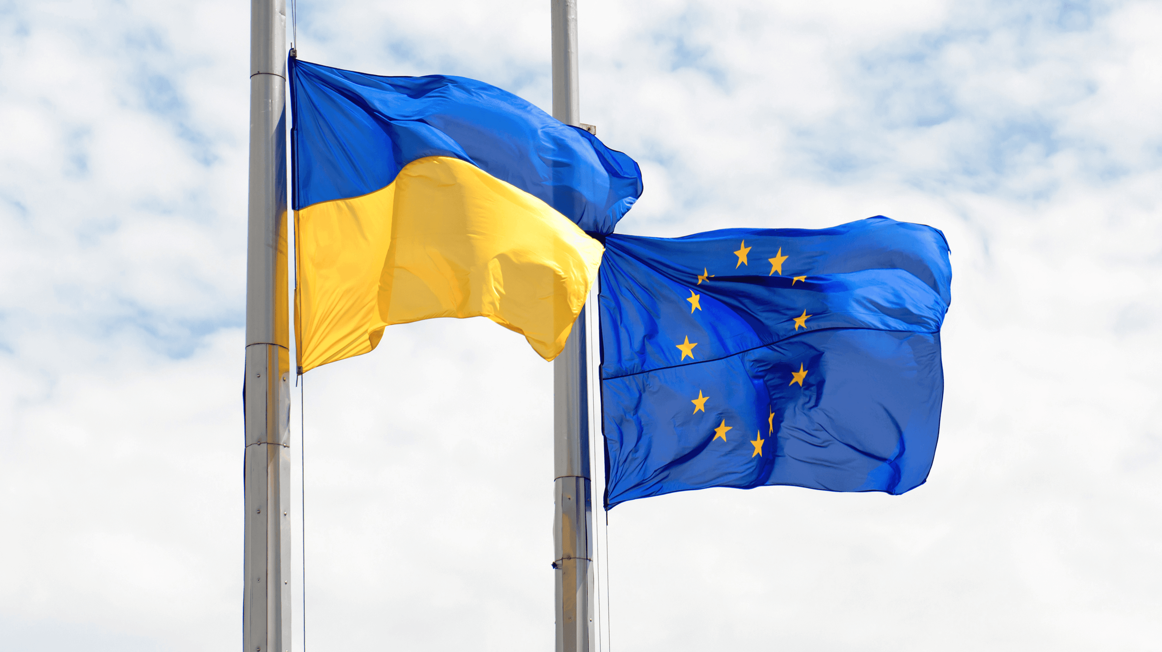 На зображенні два прапори, що майорять на флагштоках: зліва – прапор України (жовто-блакитний), а справа – прапор Європейського Союзу (синій із жовтими зірками). Фон – хмарне небо.