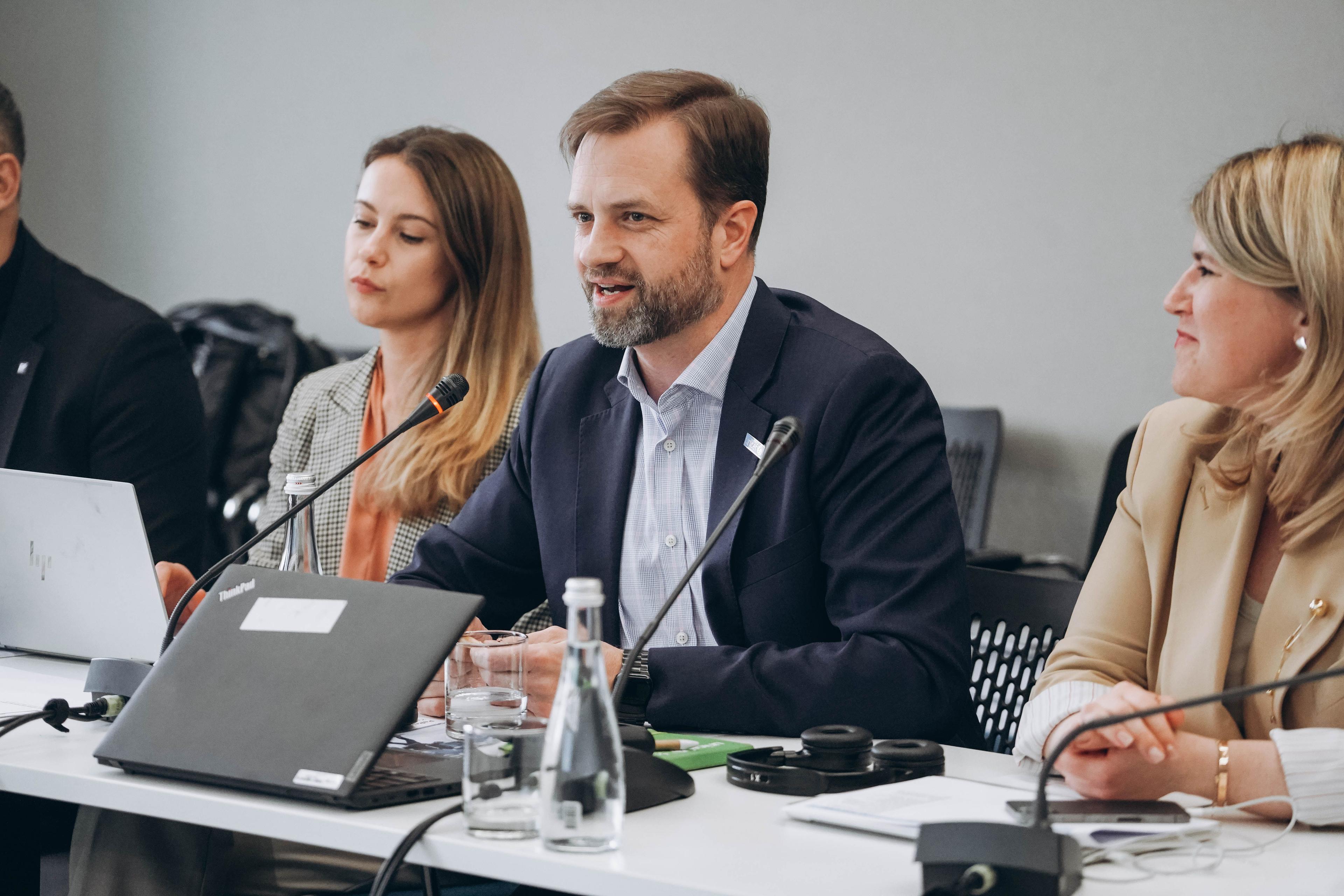 На зображенні люди на офіційній зустрічі. Перед ними ноутбуки, мікрофони. Чоловіки і жінки посміхаються