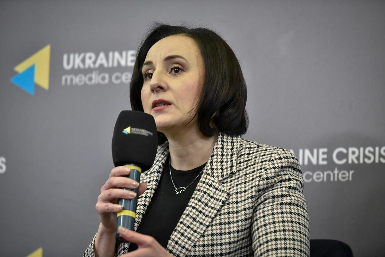 Жолнович Оксана тримає мікрофон і говорить на прес-конференції. Вона одягнена в клітчастий піджак і чорно-сірий топ, на шиї носить кольорову підвіску у вигляді карти України. На фоні видно логотипи Українського кризового медіа-центру.