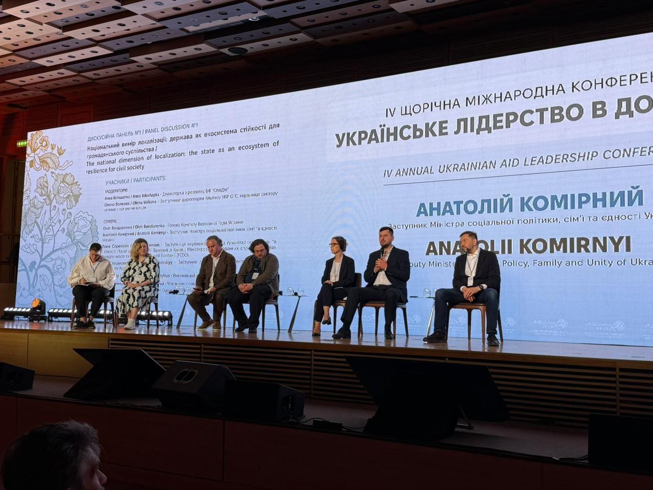 На фото чоловіки та жінки сидять на фоні банеру під час заходу