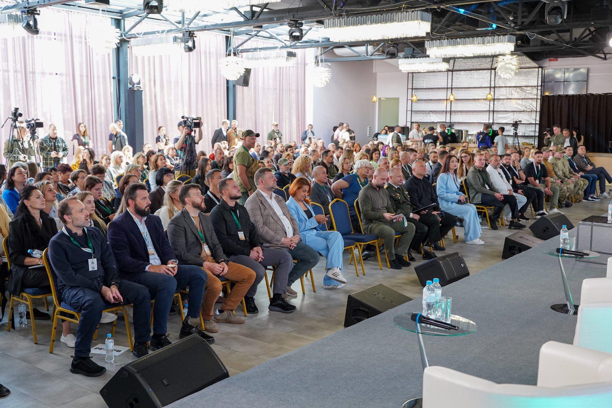 На фото чоловіки та жінки сидять у залі під час заходу