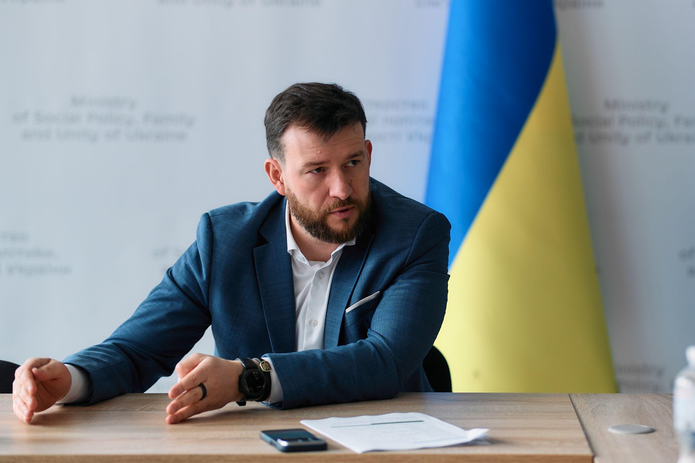 На фото чоловік сидить за столом на фоні прапору України