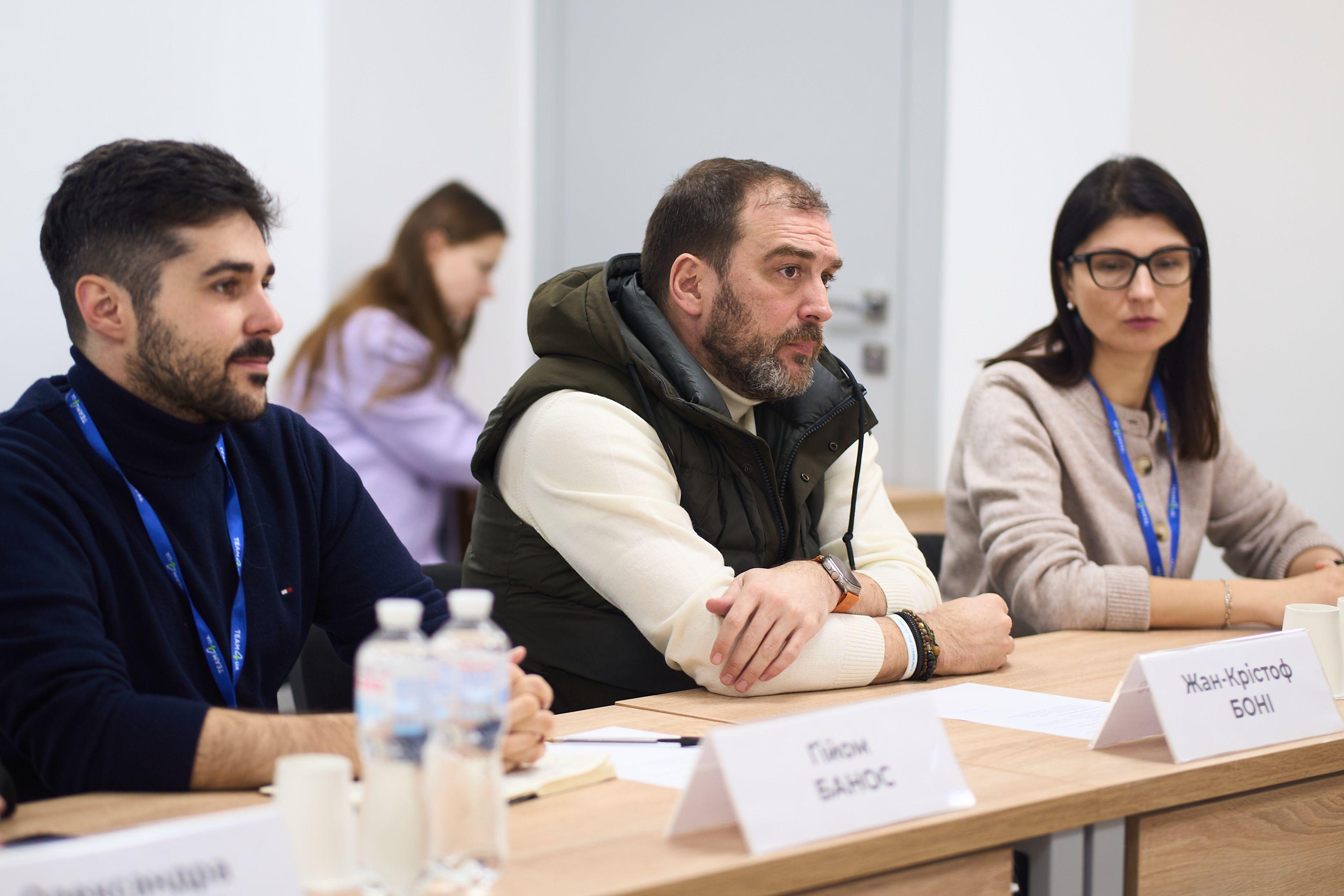 На фото чоловіки та жінка сидять за столом під час зустрічі