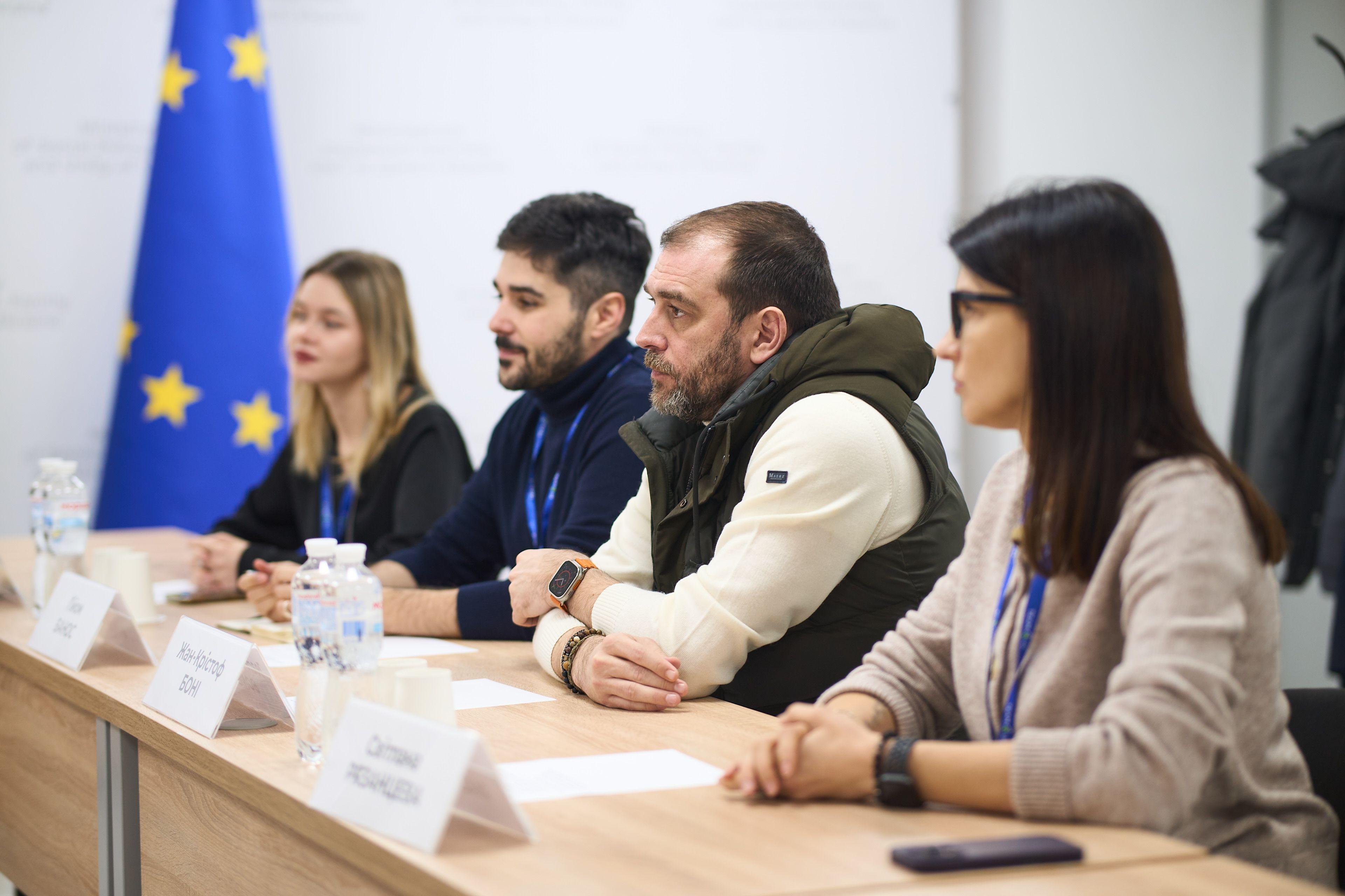 На фото чоловіки та жінки сидять за столом під час зустрічі