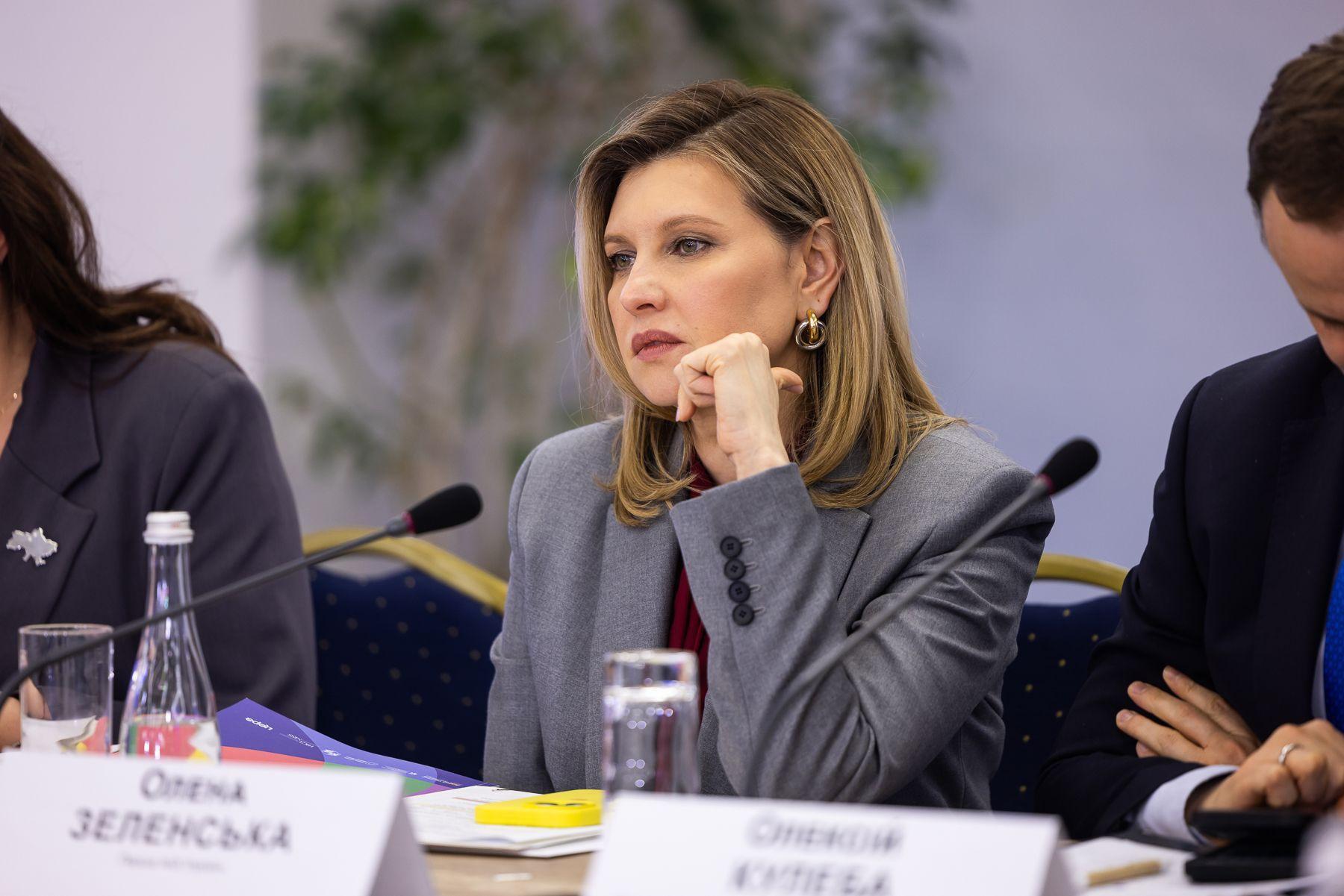 На зображенні жінка у сірому піджаку сидить за столом на офіційному заході. Вона спирається підборіддям на руку та виглядає зосередженою. Перед нею мікрофон, склянка води та документи. На табличці з ім’ям написано «Олена Зеленська».