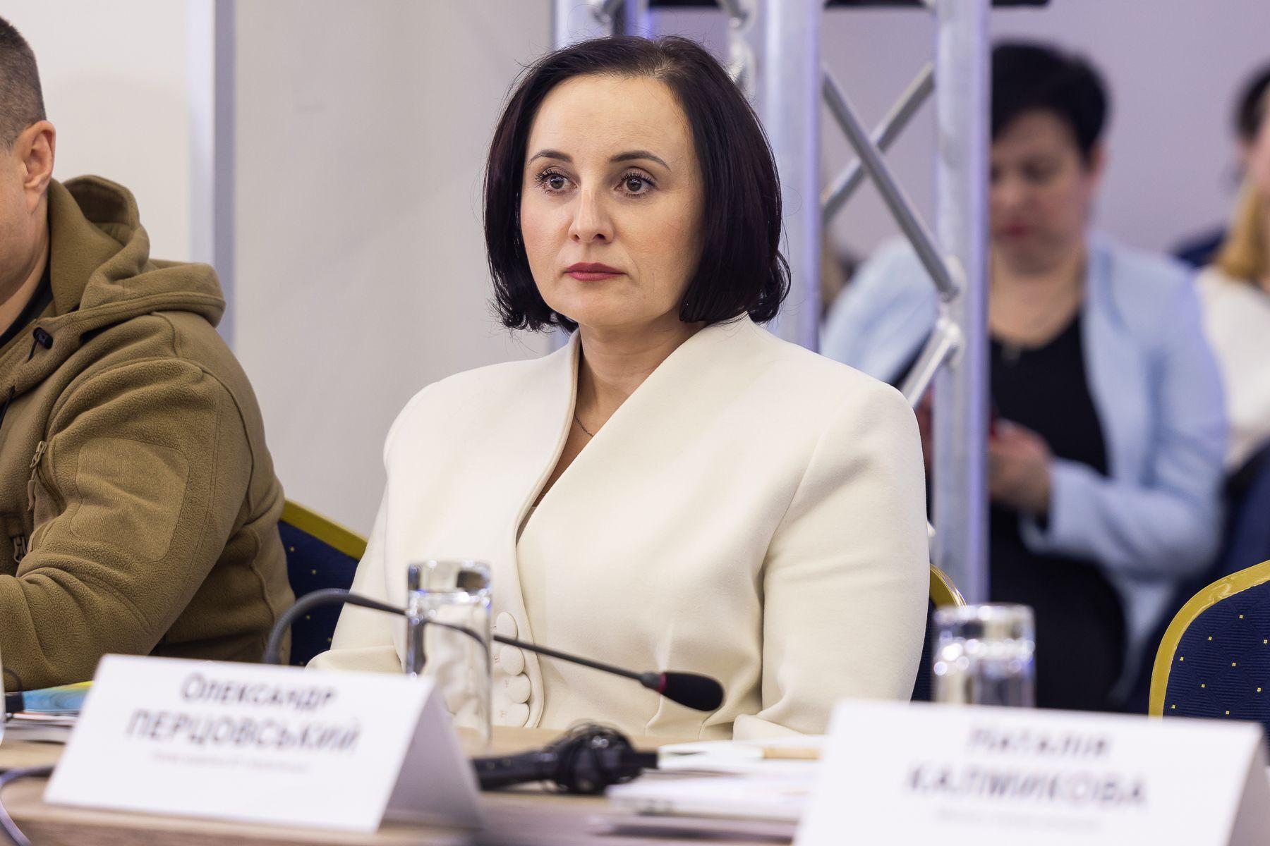 На зображенні жінка, Міністерка Оксана Жолнович, з коротким чорним волоссям у білому костюмі сидить за столом на офіційному заході. Вона має серйозний вираз обличчя. Перед нею мікрофон, склянка води та документи