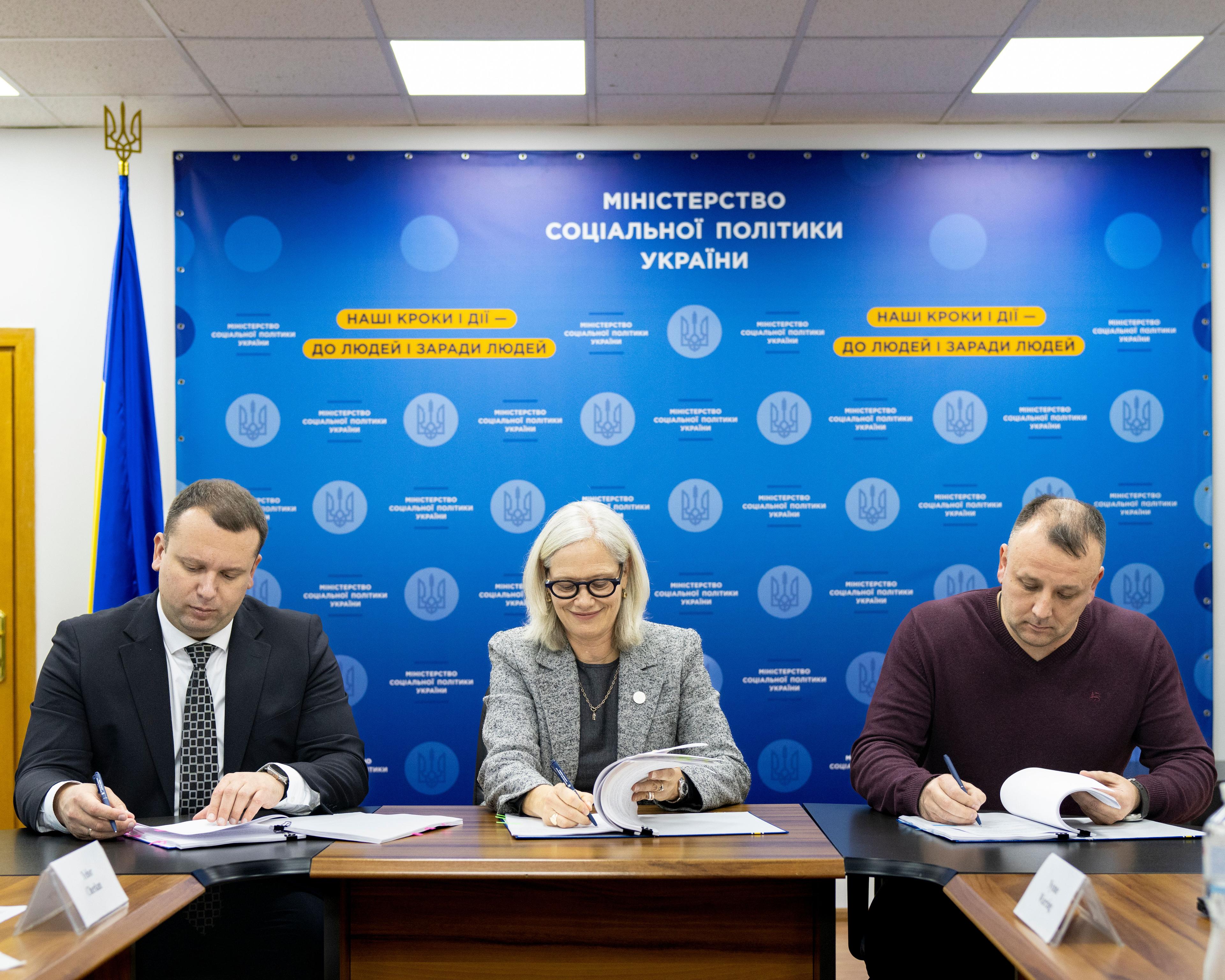 Дві особи, одна в білої сорочці, інша в темному костюмі, підписують документи на столі. Одна з них тримає ручку, а друга руку покладає на папери. На фоні видно комп'ютерну клавіатуру та годинник на руці одного з учасників.