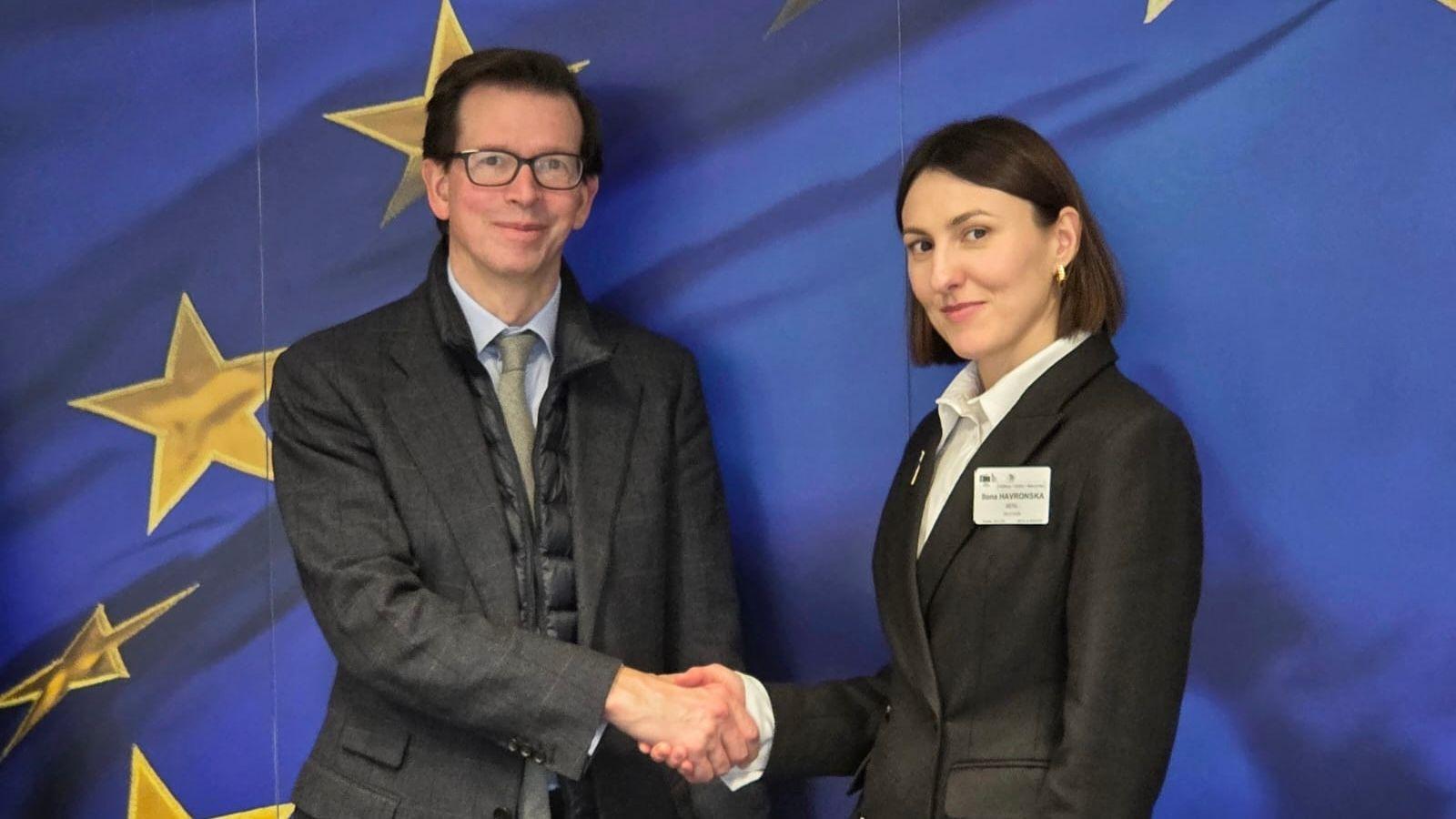 На фото чоловік і жінка вітаються за руку на фоні прапору ЄС