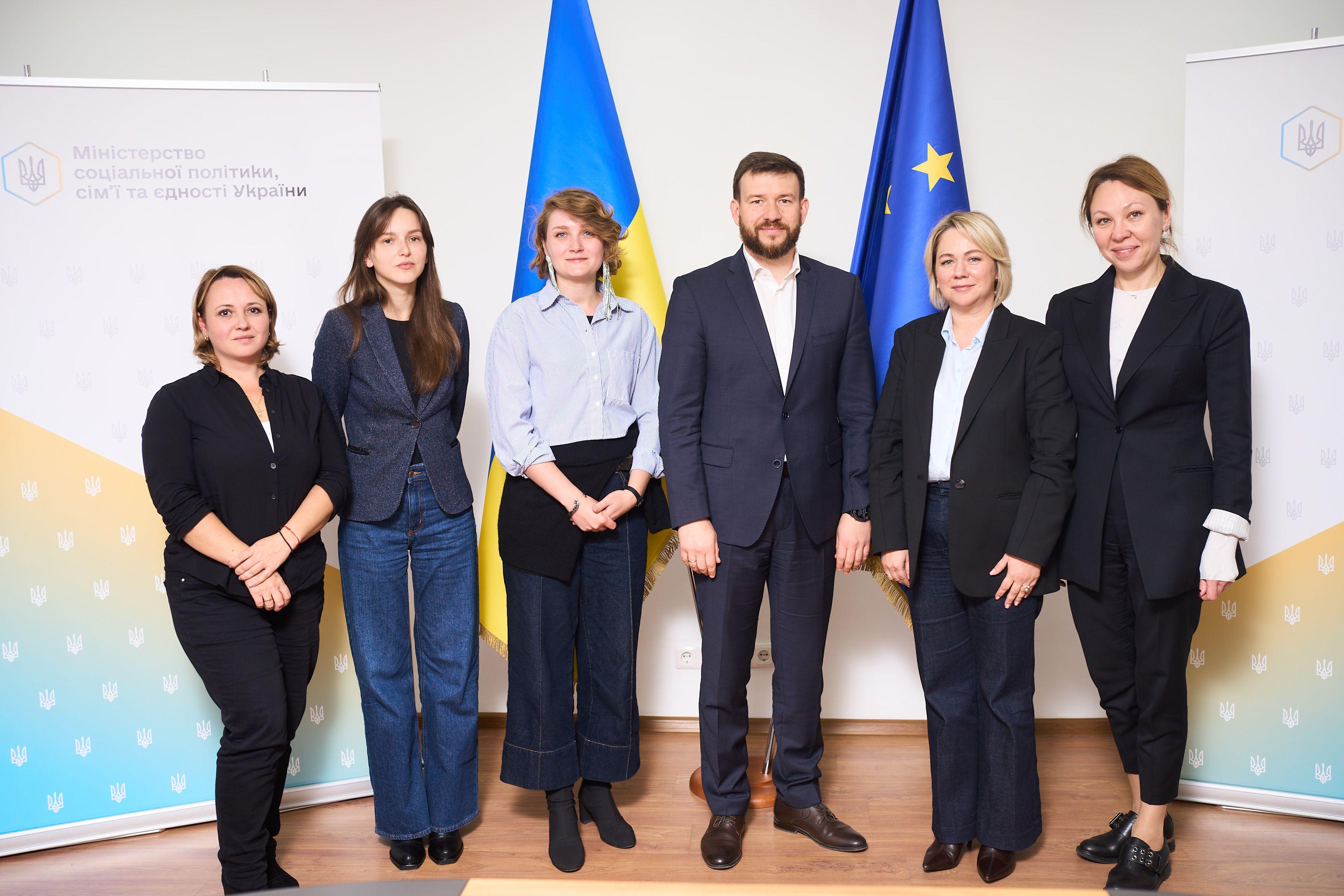 НА фото єінки та чоловік стоять на фоні прапорів України та ЄС