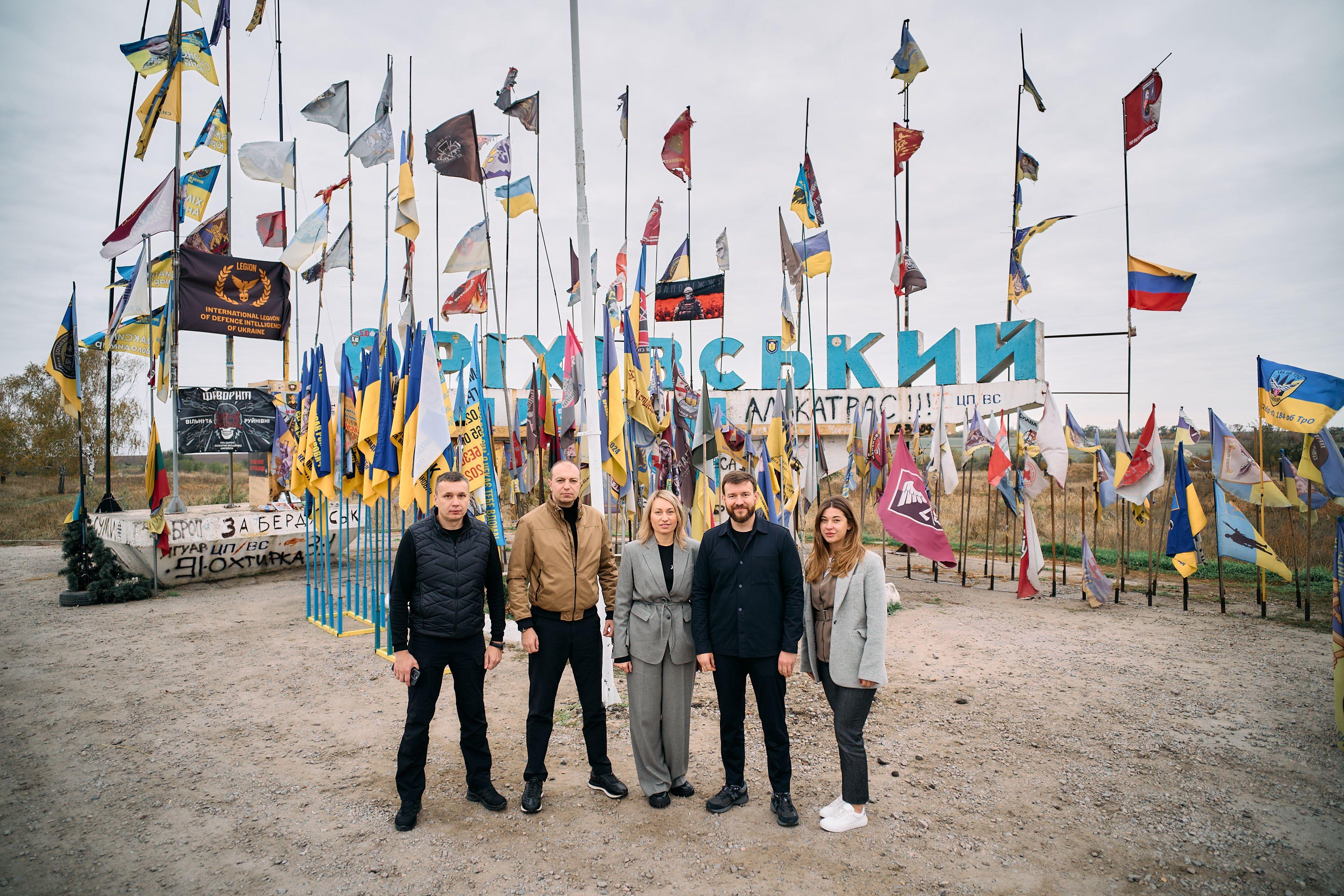 На фото чоловіки та жінка стоять на фоні прапорів