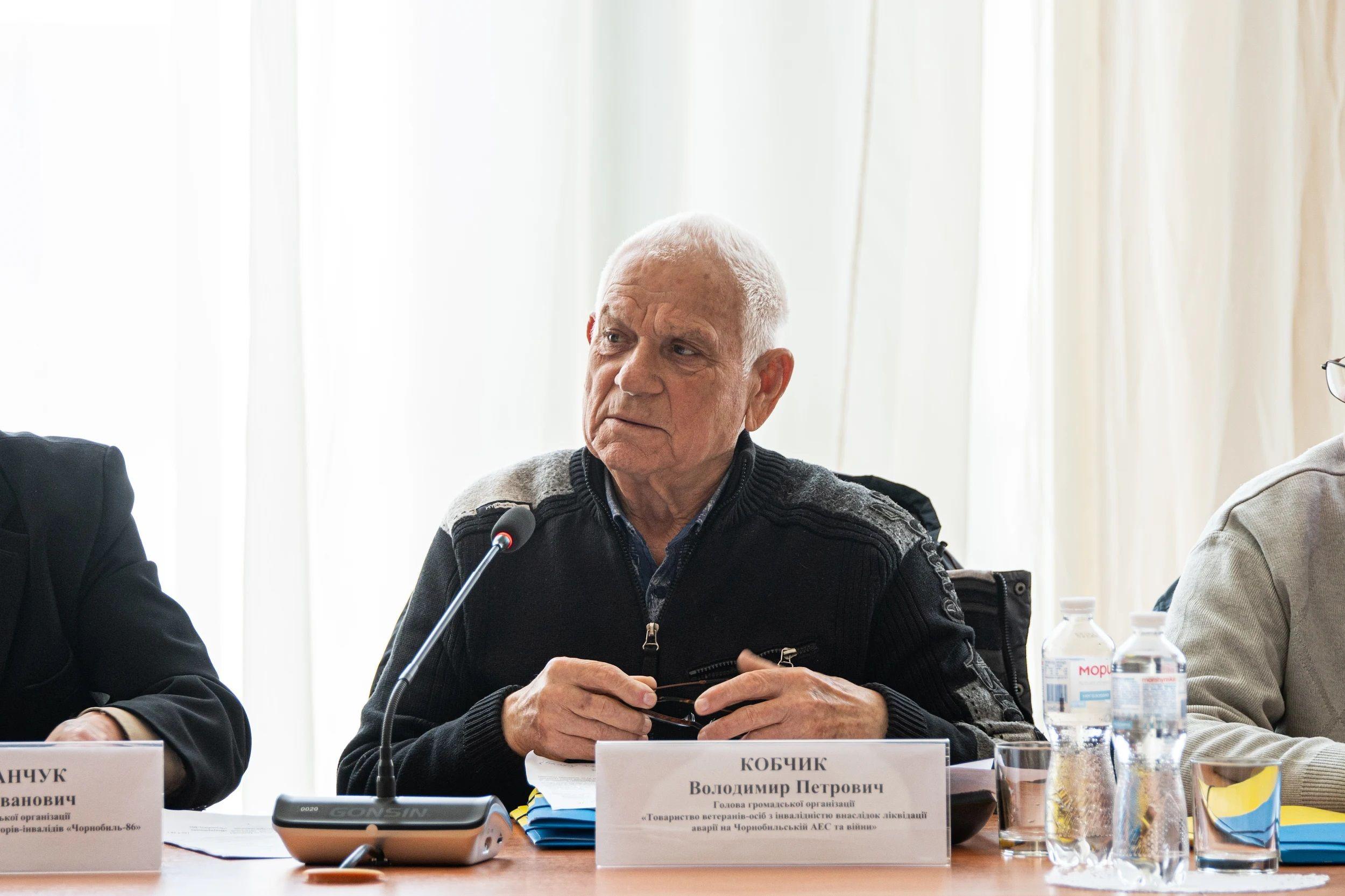 На фото чоловік в окулярах сидить за столом під час слухань