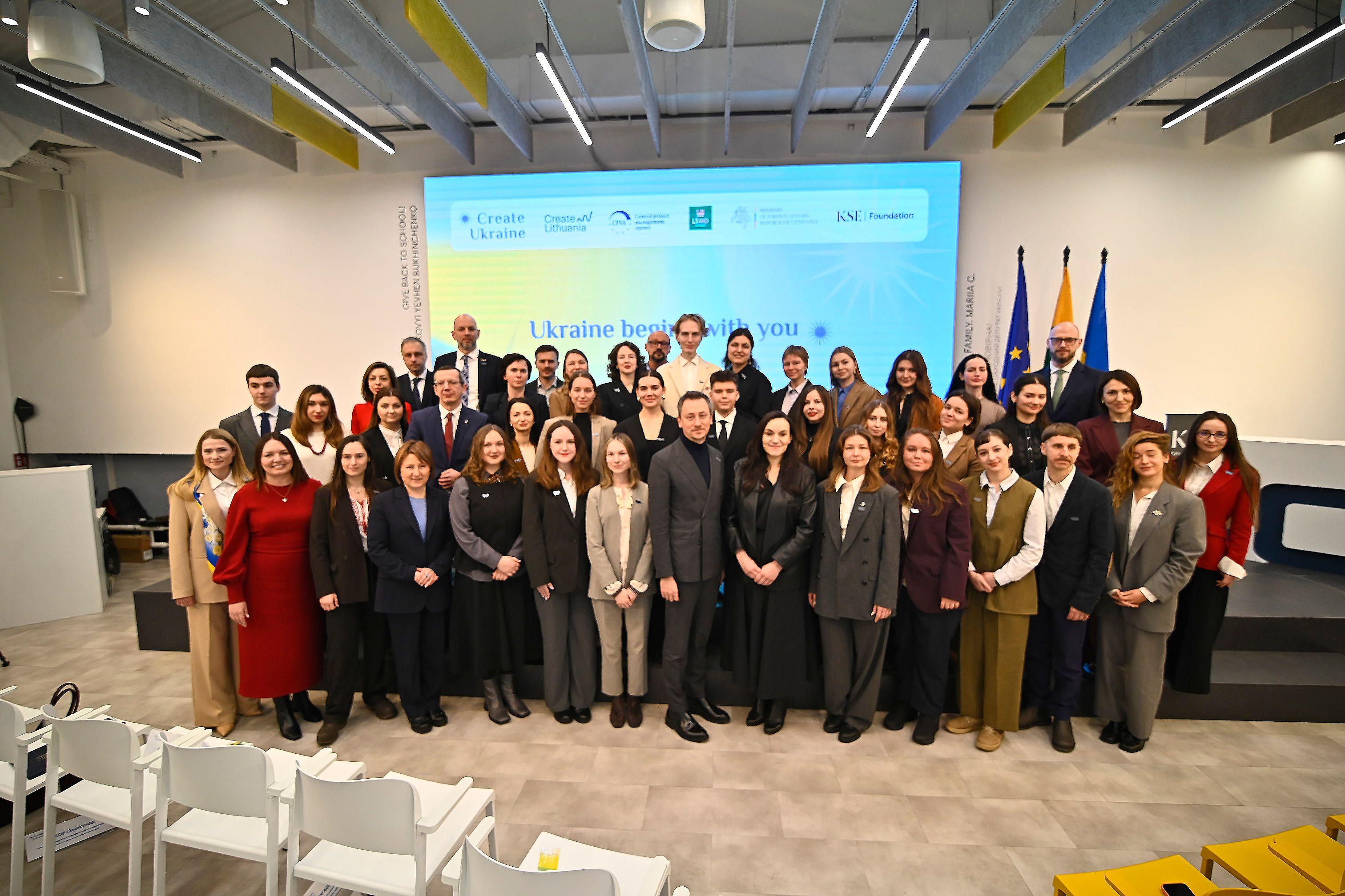 На фото жінки та чоловіки стоять на фоні сцени та прапорів