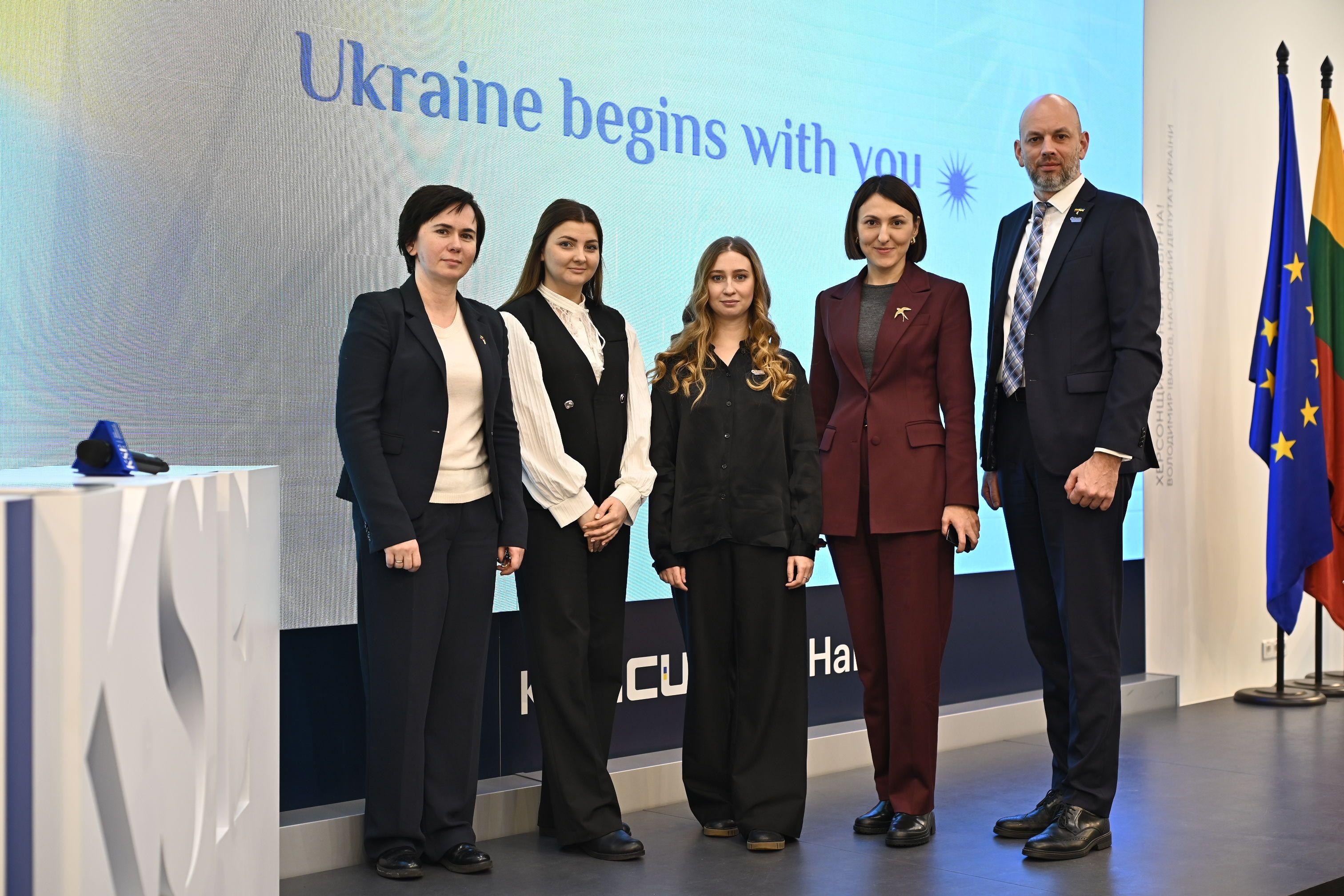 На фото жінки та чоловік стоять на фоні банеру