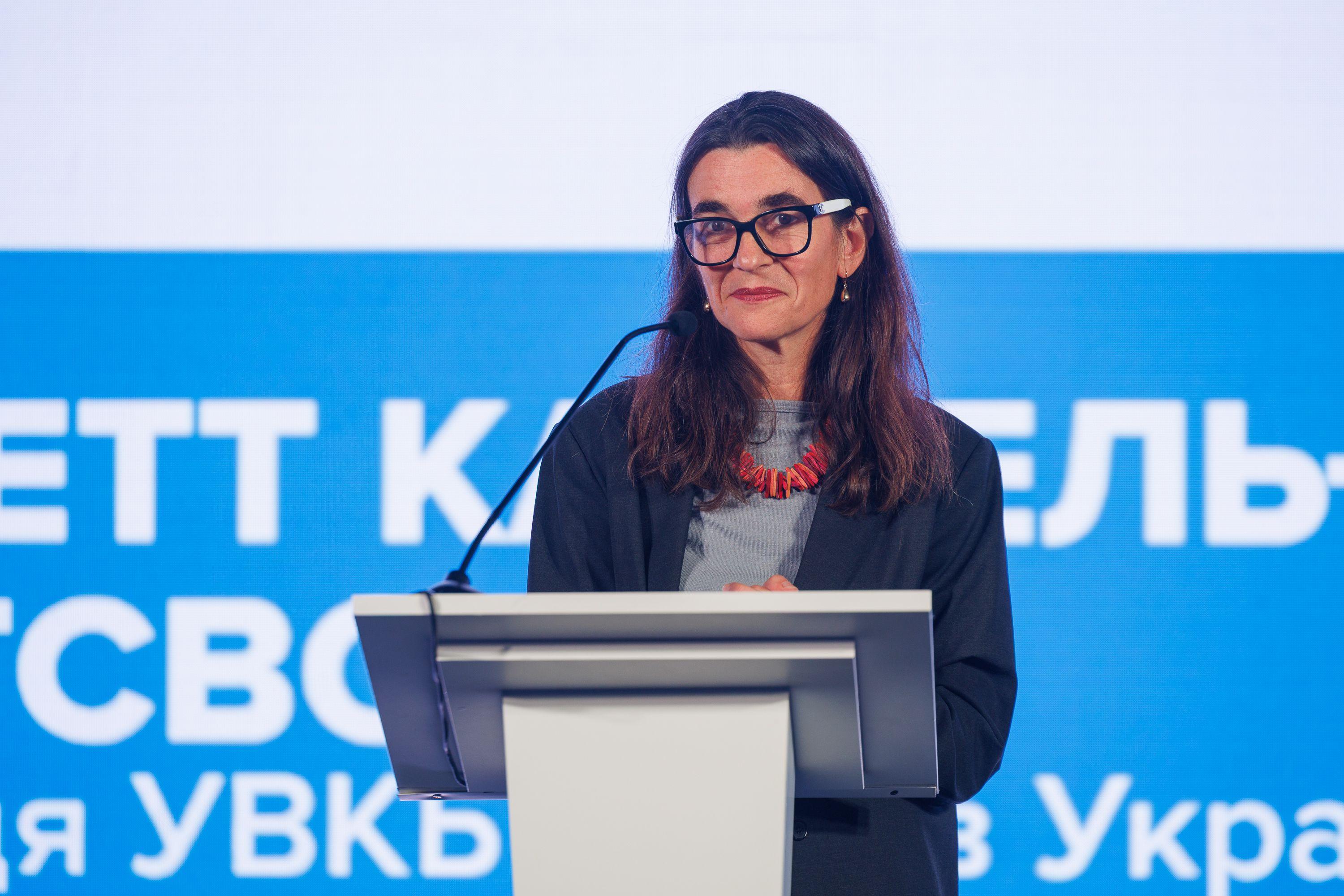 На фото жінка в окулярах виступає перед учасниками Форуму