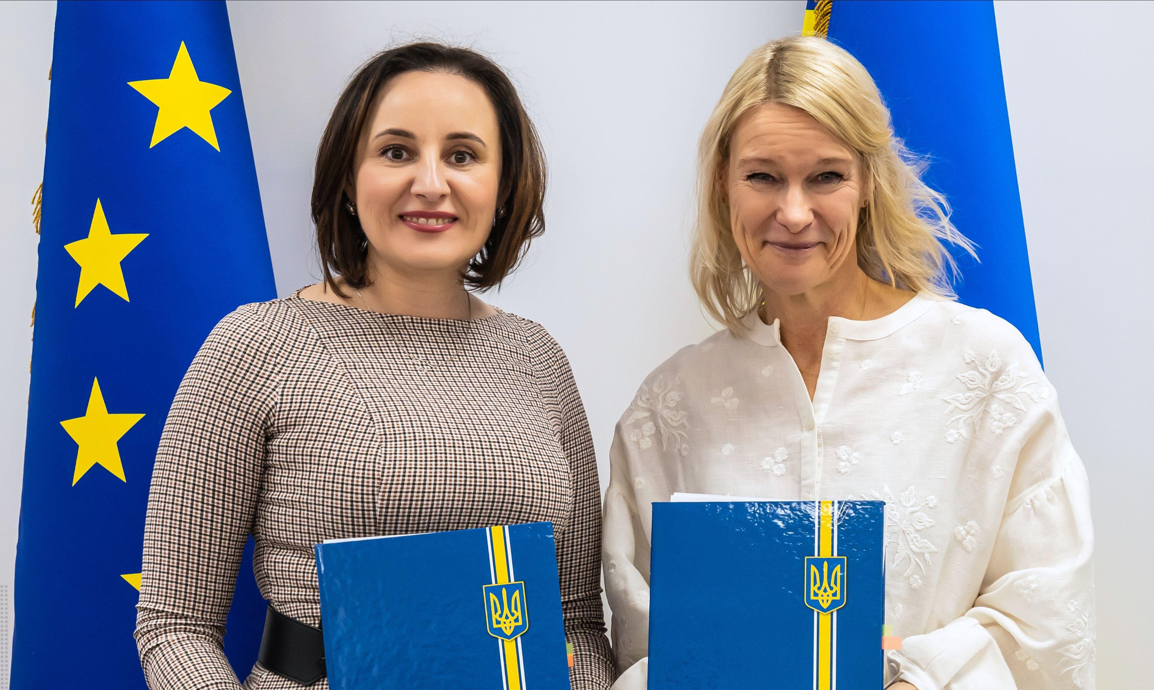 На зображенні дві жінки посміхаються і тримають в руках папки. Одна з ним Міністерка соціальної політики Оксана Жолнович