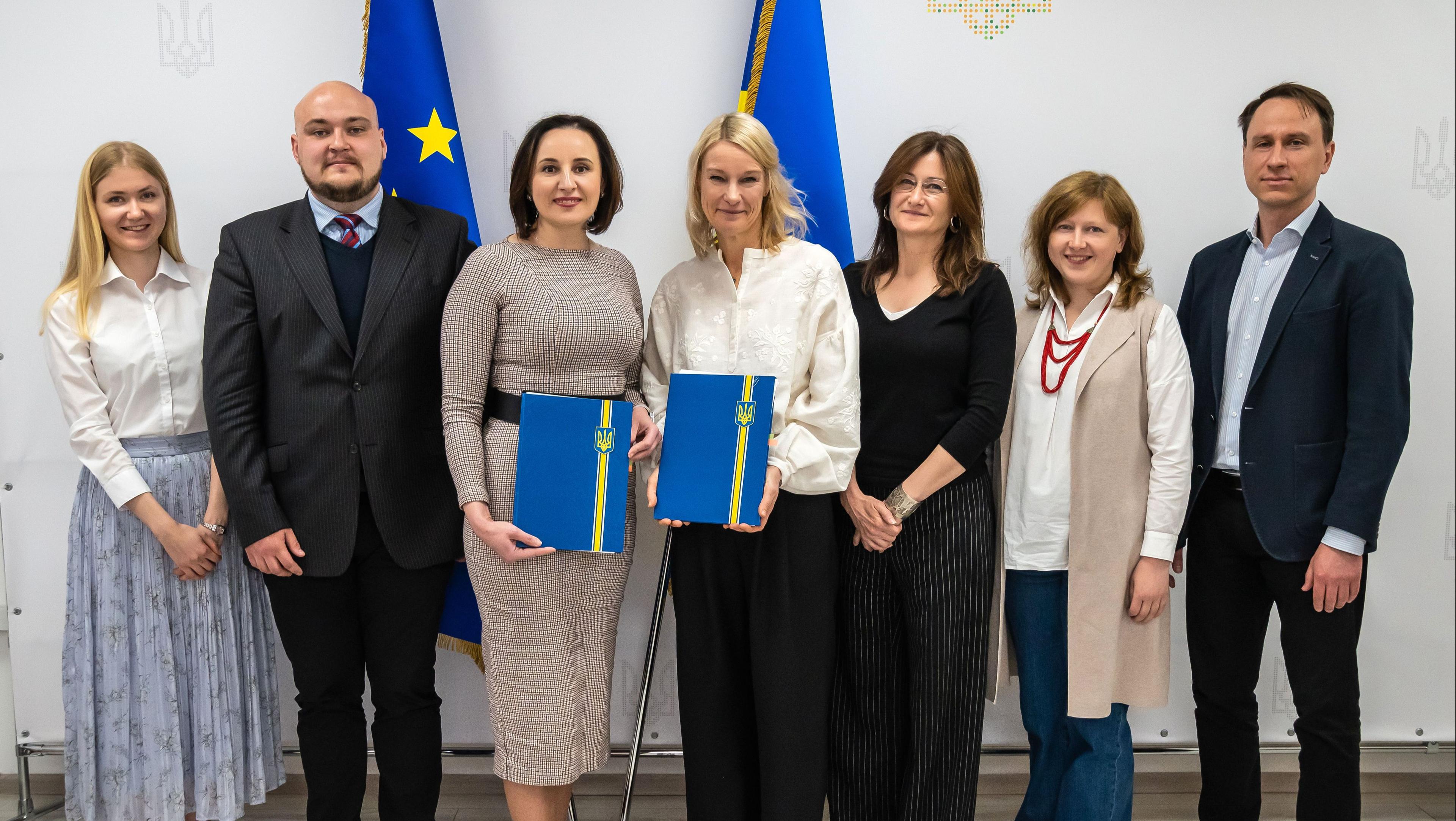 На фото група людей після підписання документів. Посміхаються і тримають в руках папки синього кольору. 