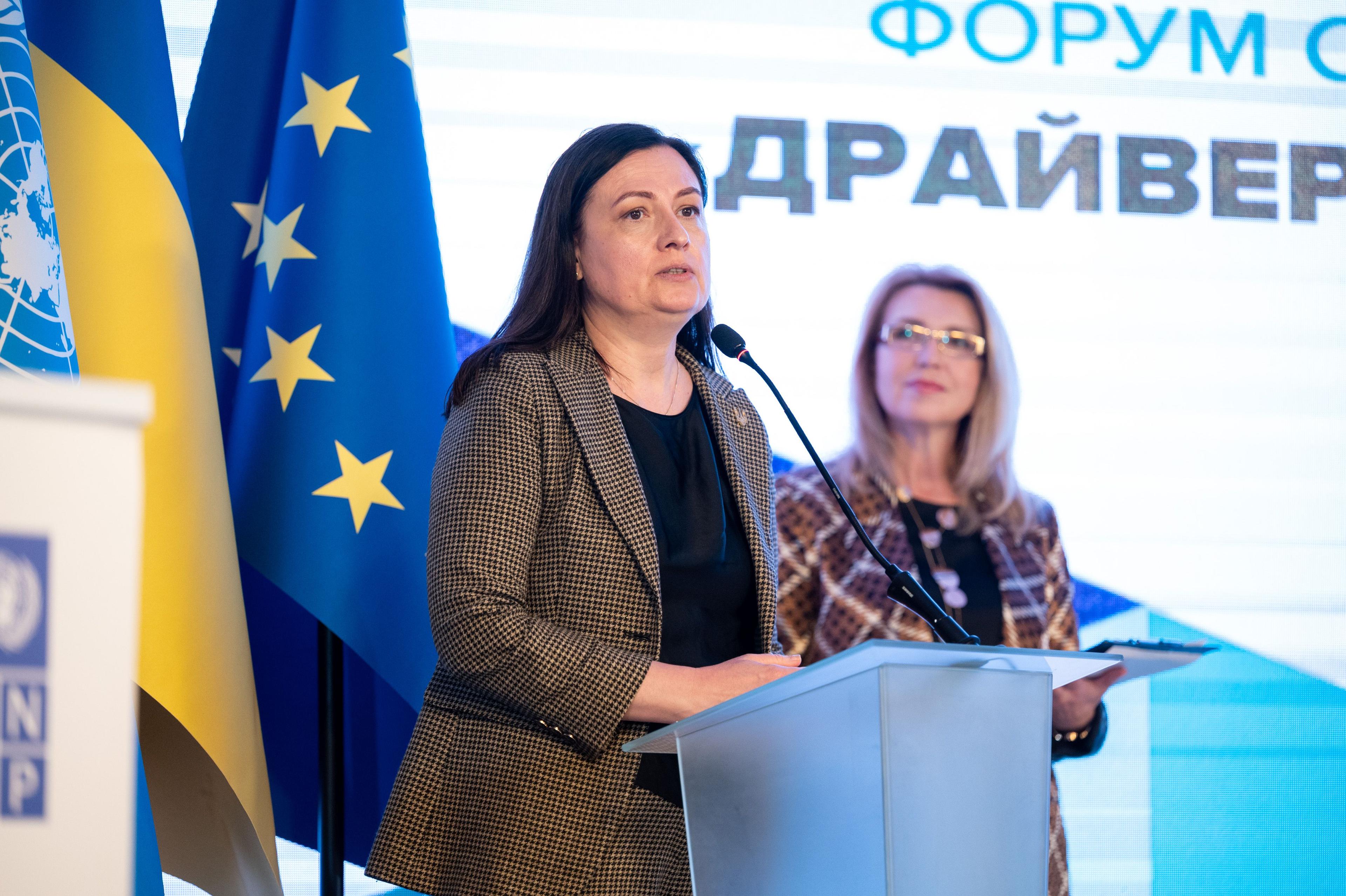 На зображенні жінка, Тетяна Ломакіна, за трибуною з мікрофоном щось доповідає
