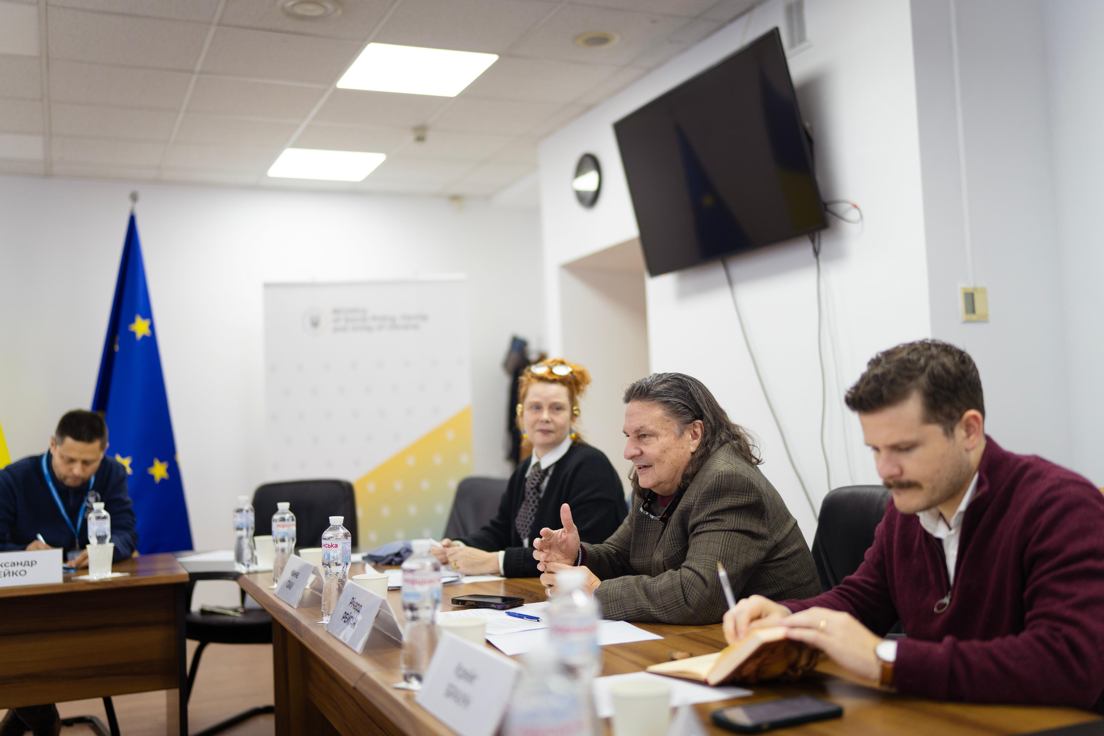 На фото жінка та чоловіки сидять за столом під час зустрічі