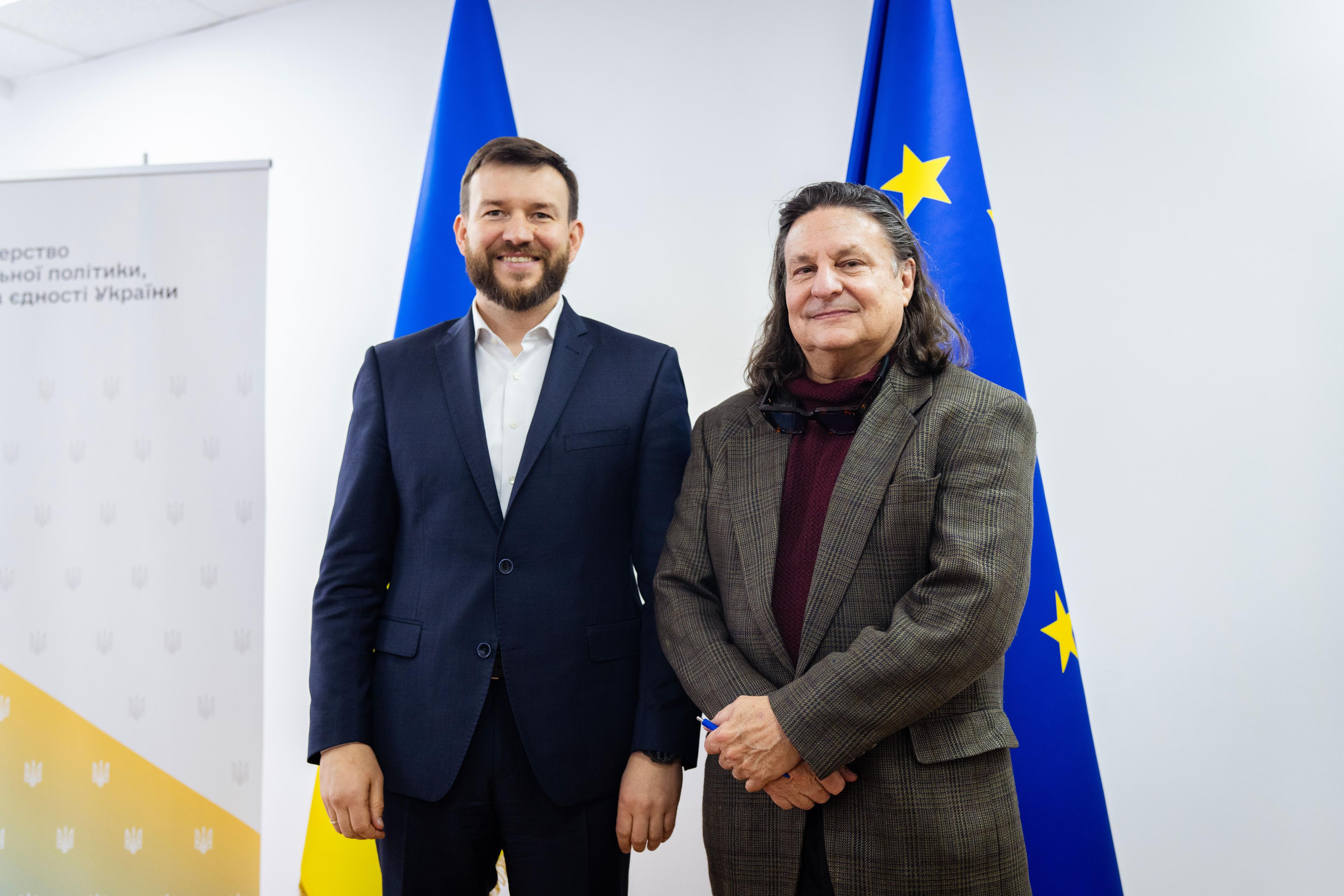 На фото двоє чоловіків стоять на фоні прапорів України та  ЄС