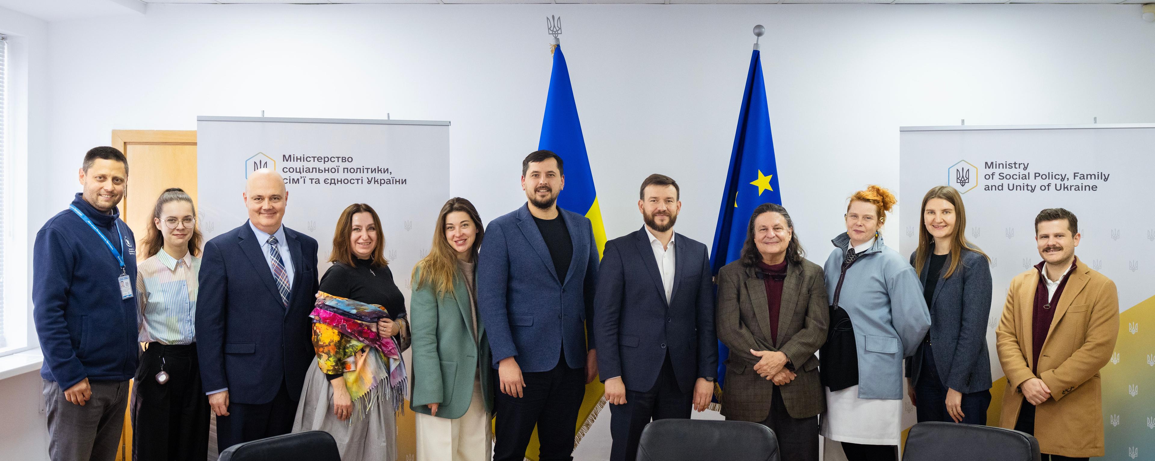 На фото жінки та чоловіки стоять на фоні прапорів України та ЄС