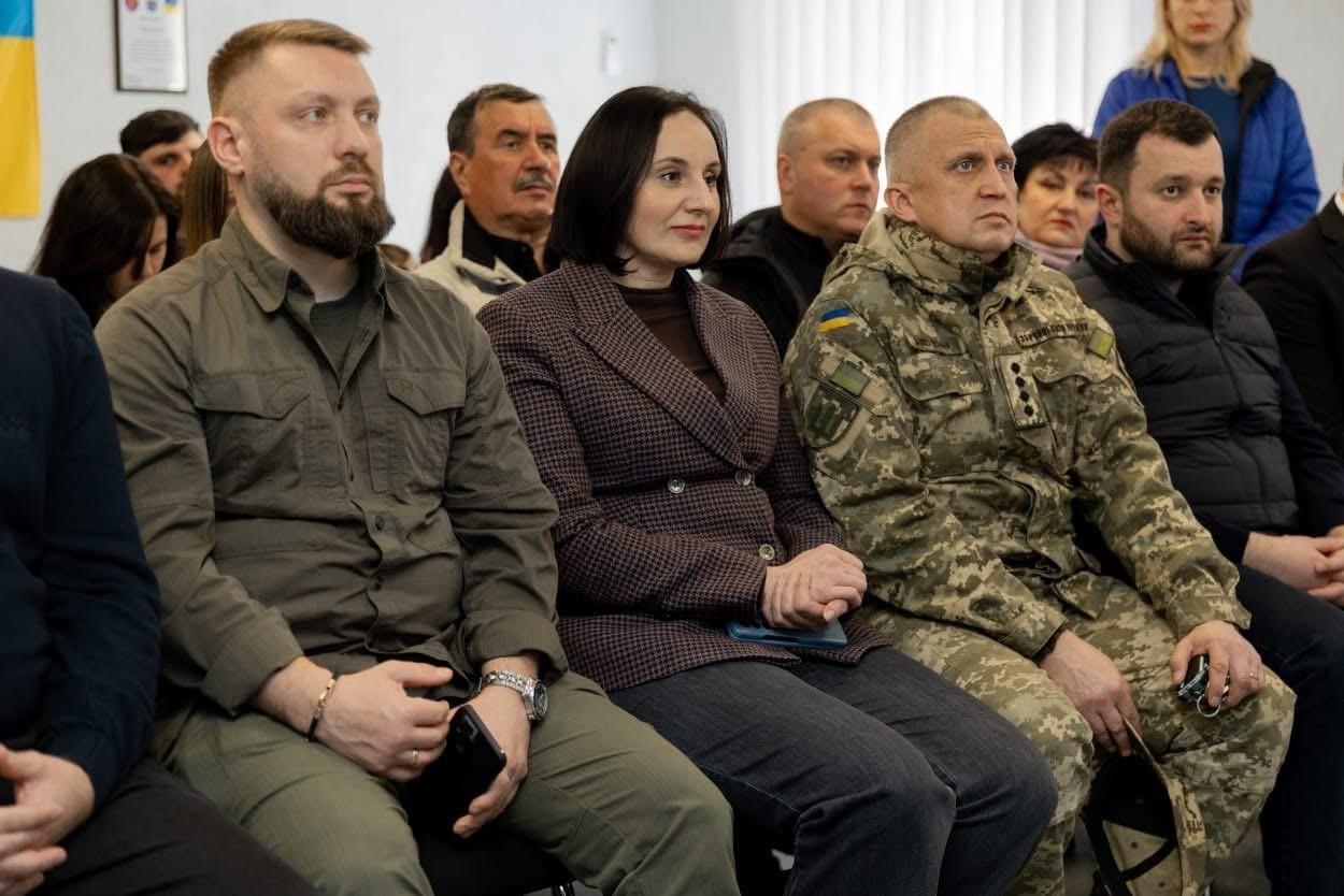 На зображенні група людей сидить у приміщенні, уважно слухаючи щось або когось. У центрі – жінка в піджаку з коричневим візерунком, поруч з нею чоловік у військовій формі з українським шевроном на рукаві. Ліворуч від жінки – чоловік у оливковій сорочці.