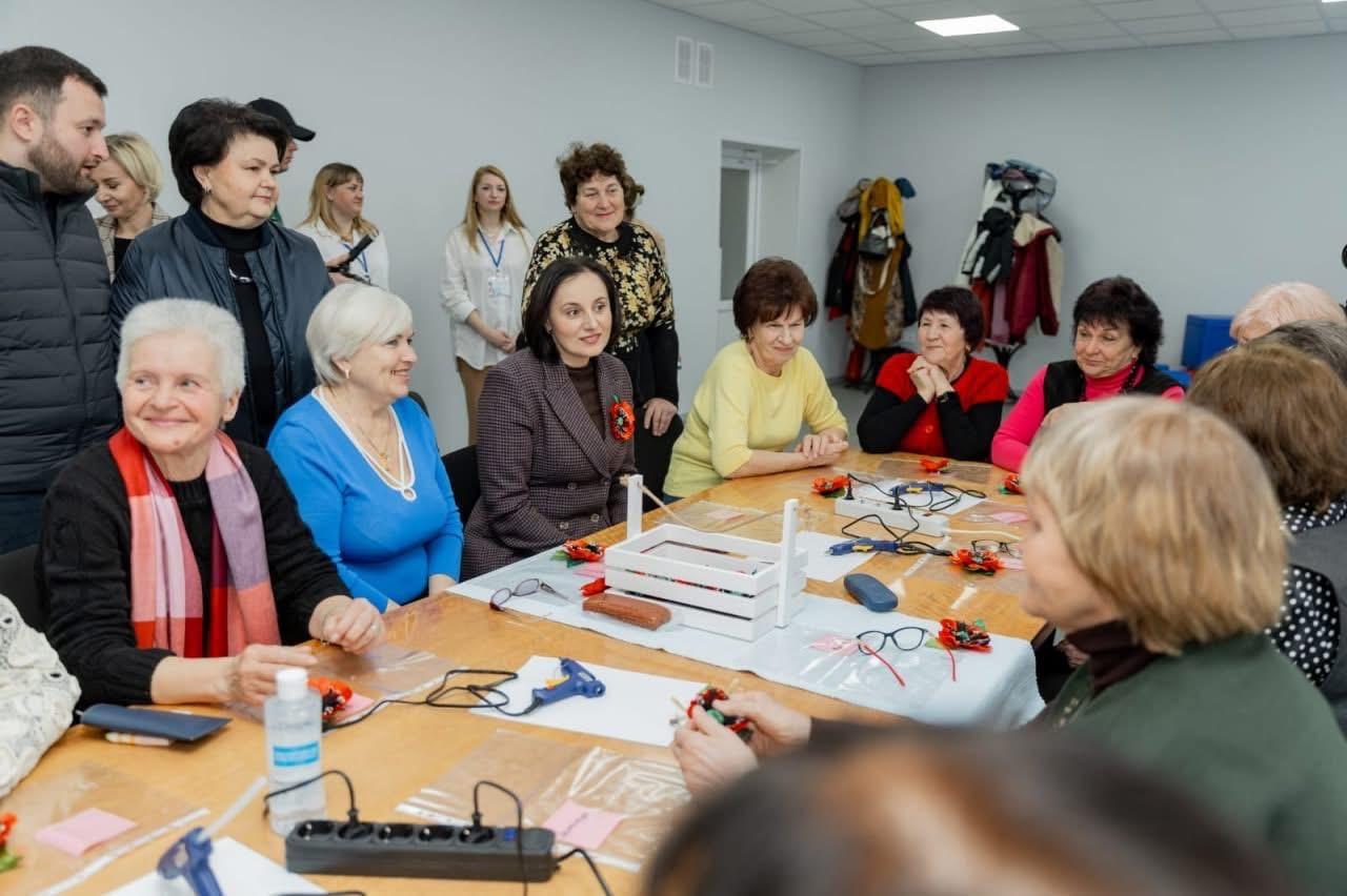 На зображенні група людей, переважно жінок, бере участь у творчому майстер-класі. Вони сидять за столом, на якому розкладені матеріали для рукоділля, клейові пістолети, ножиці та декоративні елементи.