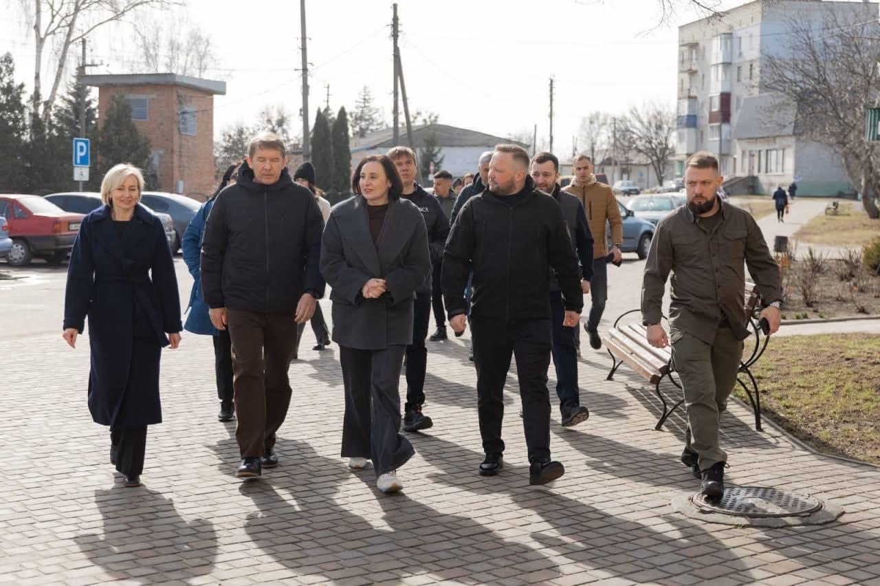 Група людей, які йдуть вулицею. Одягнені у формальний і напівформальний одяг. Довкола міське середовище з житловими будинками, автомобілями та лавками.