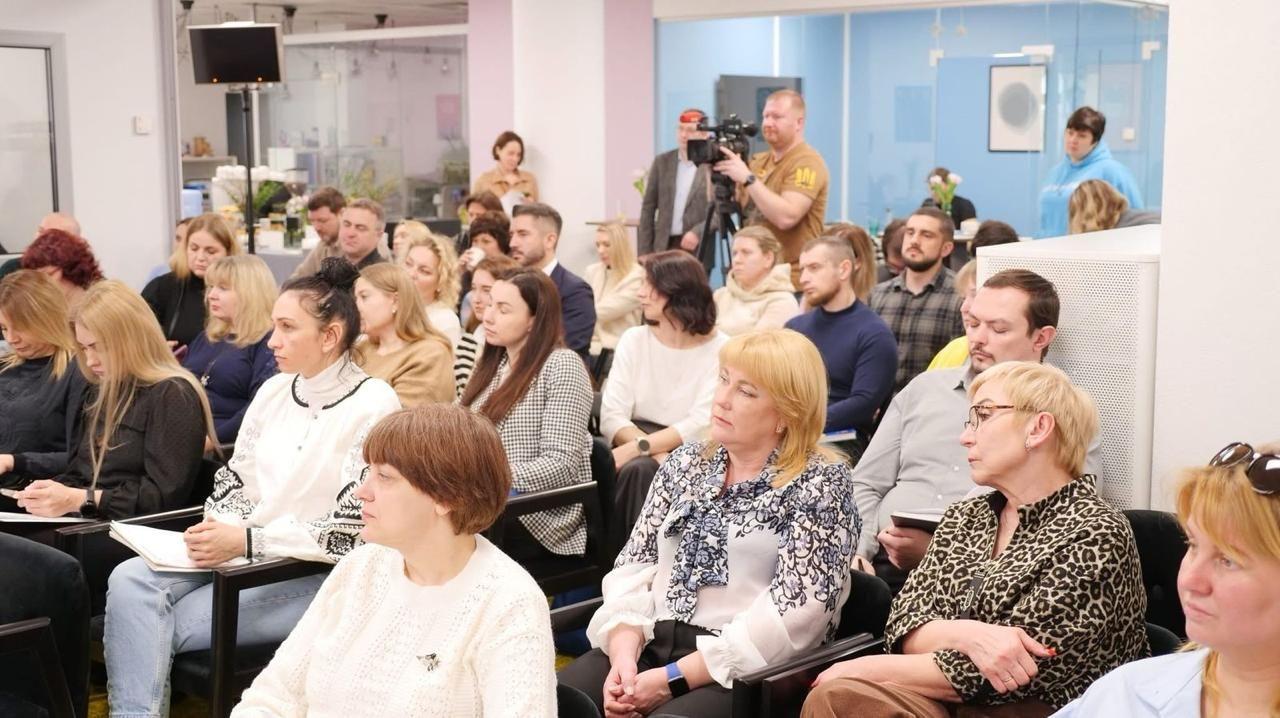 На зображенні аудиторія на публічному заході. Люди уважно слухають, деякі роблять нотатки, а інші тримають телефони. У задній частині приміщення оператор знімає подію на камеру. Приміщення світле, з сучасним дизайном та скляними перегородками