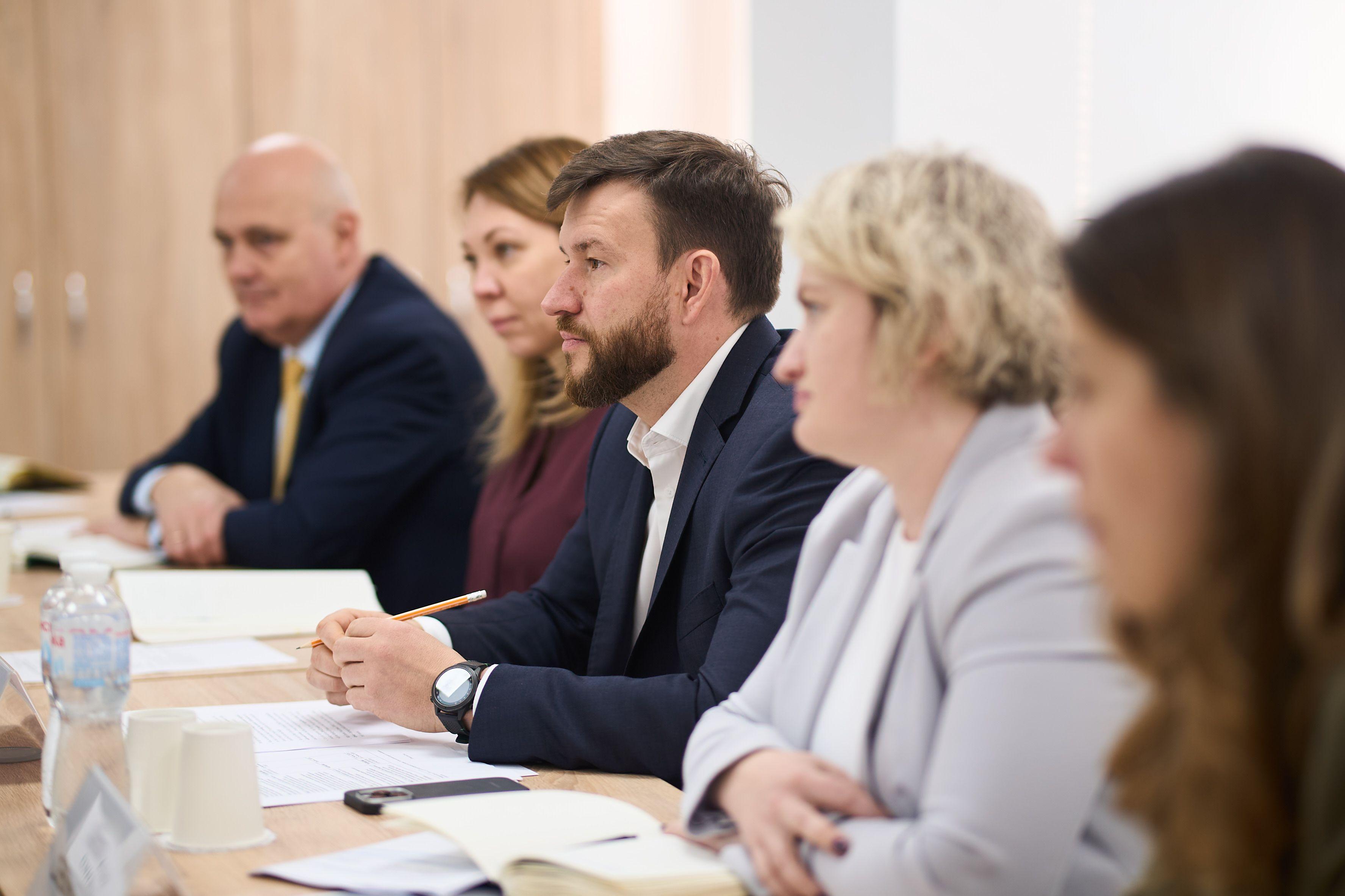 На фото чоловіки та жінки сидять за столом під час зустрічі На фото чоловіки та жінки сидять за столом під час зустрічі