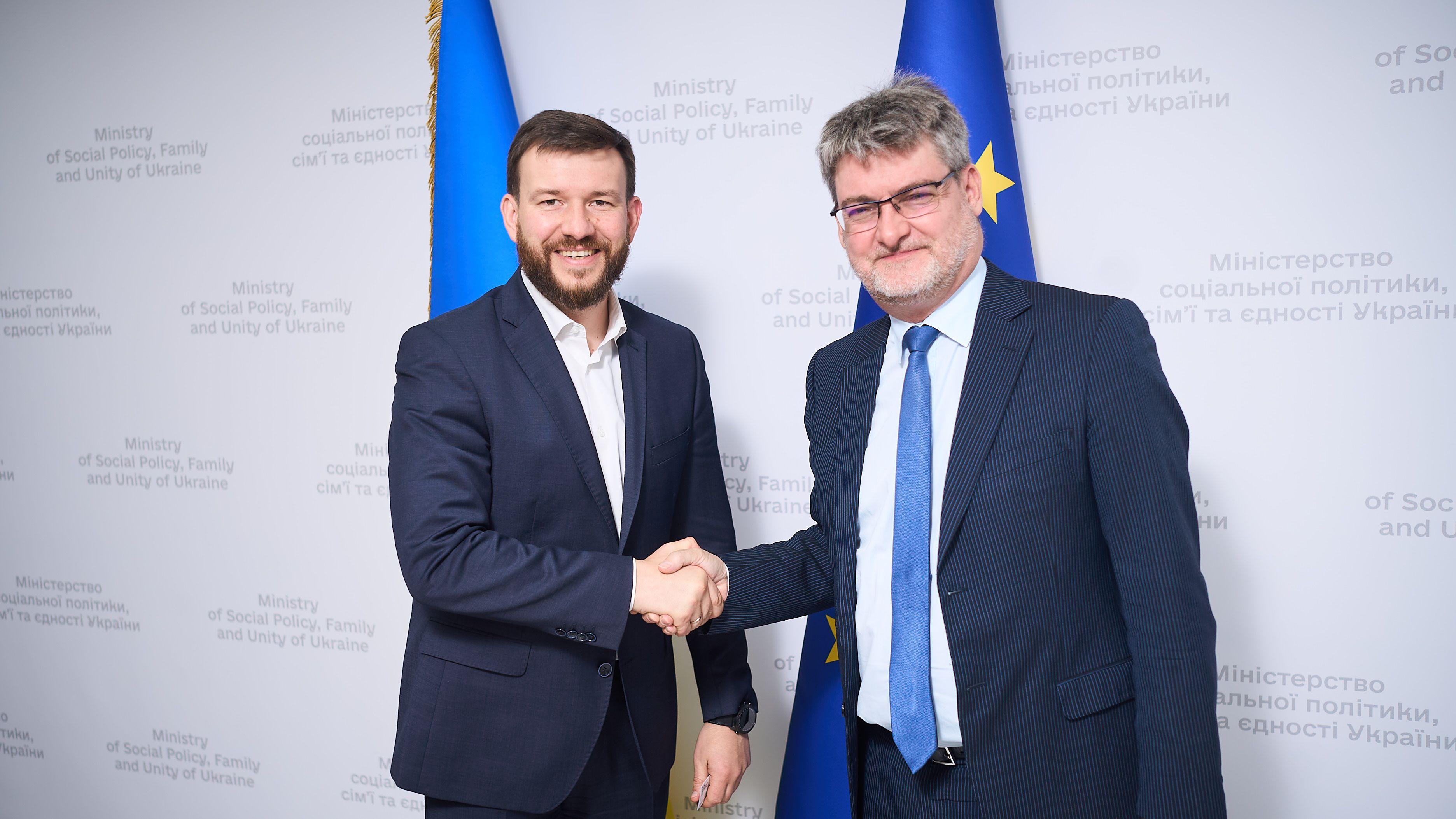 На фото чоловіки вітаються за руку на фоні прапорів України та ЄС На фото чоловіки вітаються за руку на фоні прапорів України та ЄС