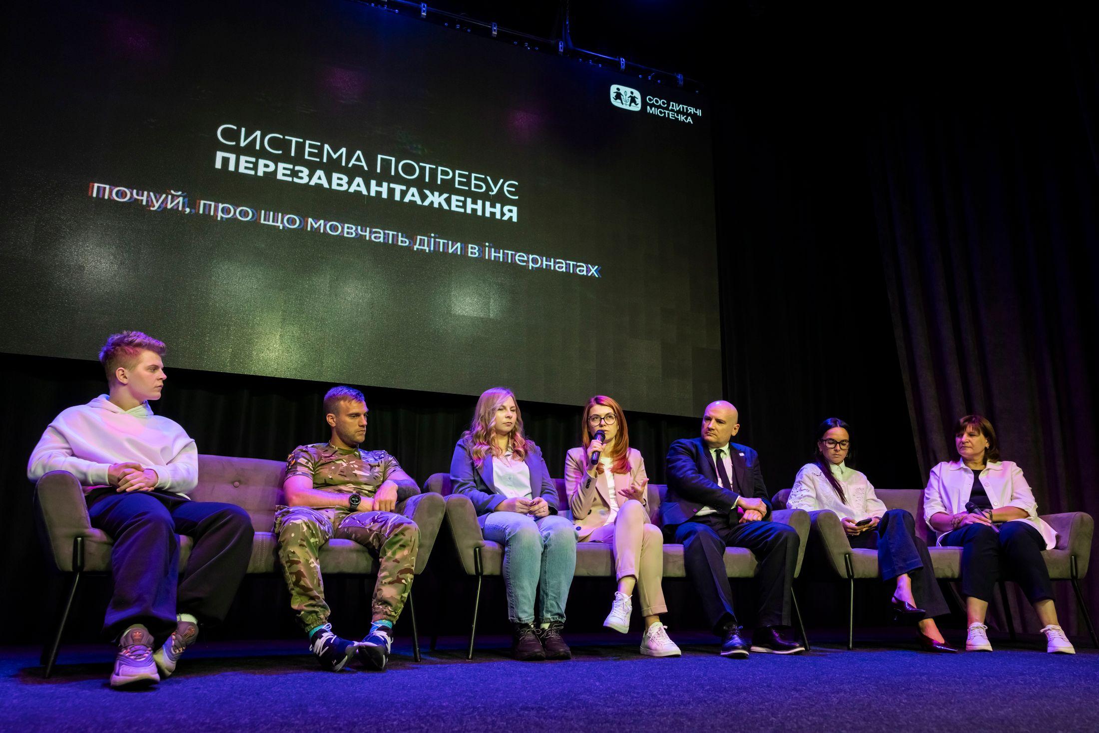 На фото чоловіки та жінки сидять в президії під час заходу На фото чоловіки та жінки сидять в президії під час заходу